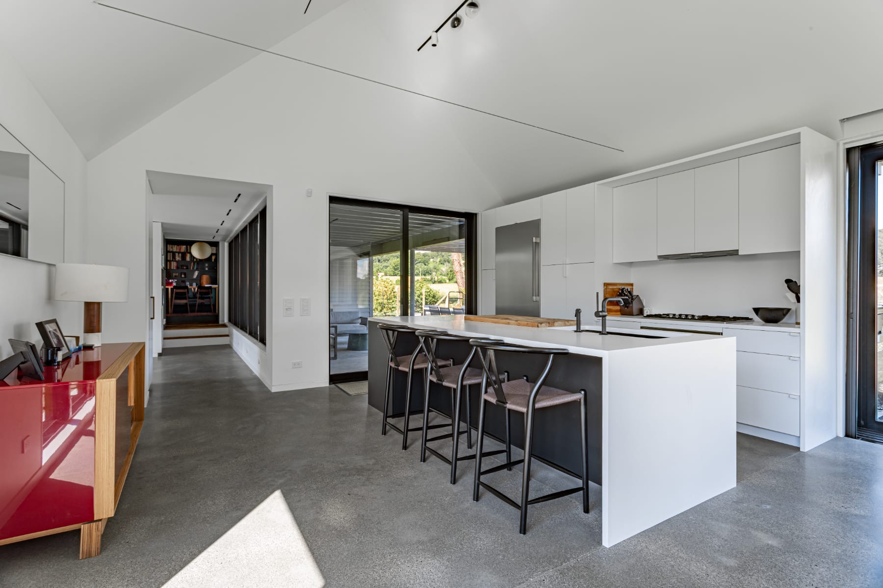 Sleek, modern kitchen featuring white cabinetry, a large island with a white countertop and black bar stools, and stainless steel appliances. Polished concrete floors and a vaulted ceiling contribute to the open, contemporary feel.