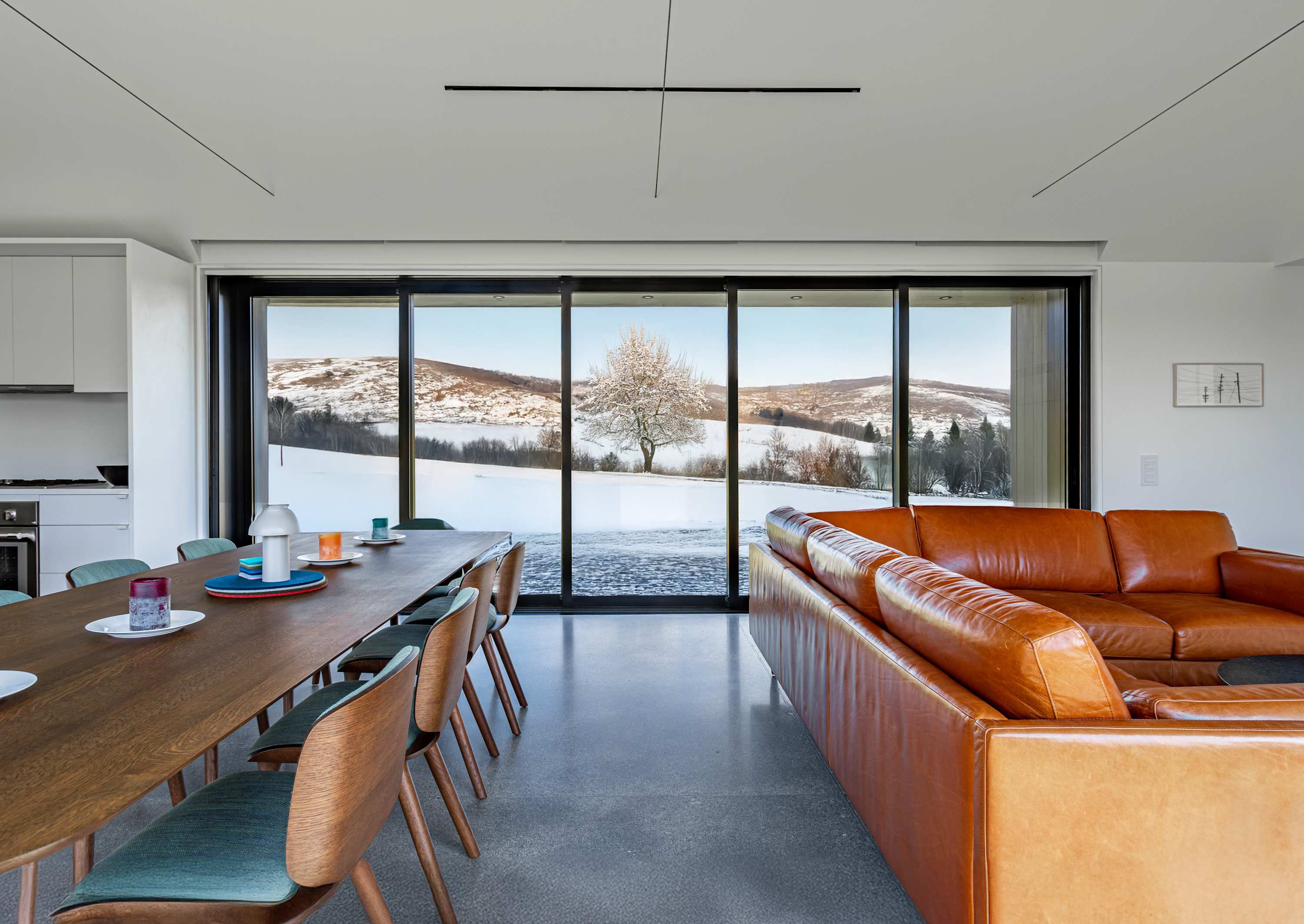 Dining area with a wooden table and chairs, offering a stunning winter view of snow-covered rolling hills and a lone tree through a large floor-to-ceiling window. The space features polished concrete floors and a modern aesthetic.