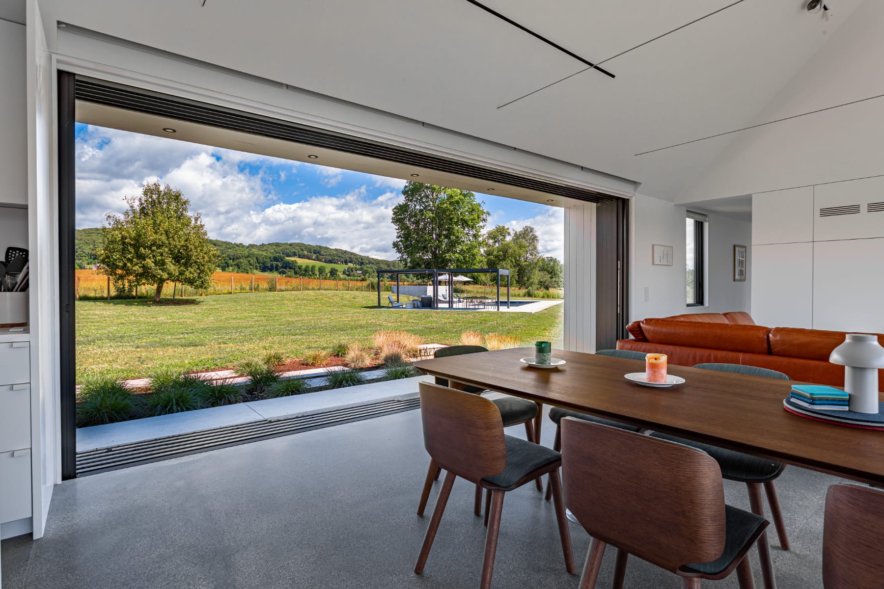 Dining area with a wooden table and chairs, featuring a large sliding glass door that opens to a lush green backyard with a distant view of the pool and surrounding landscape. The room has polished concrete floors and a modern design.