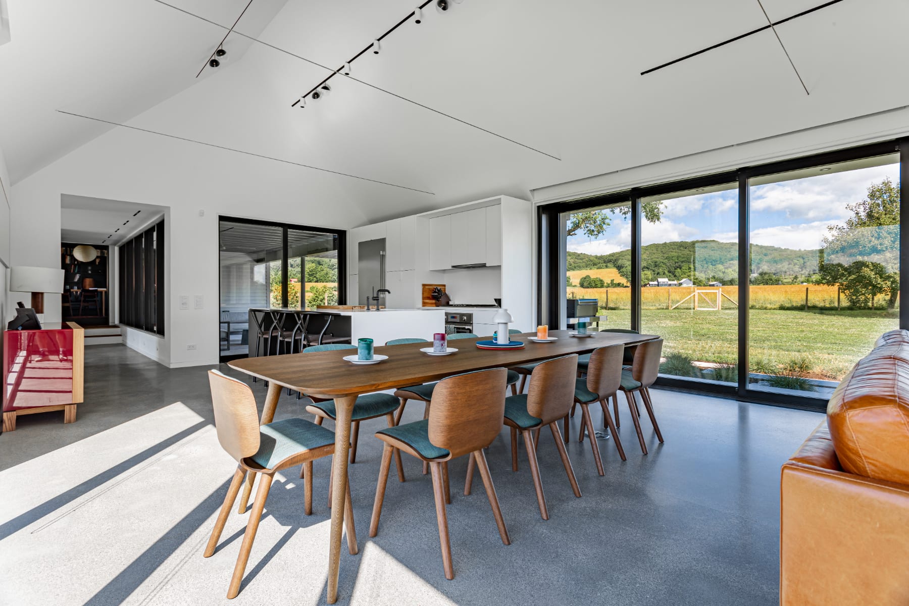 Open-concept dining area with a long wooden table and eight chairs, adjacent to a modern kitchen and living space. Floor-to-ceiling windows offer expansive views of the green outdoor landscape.