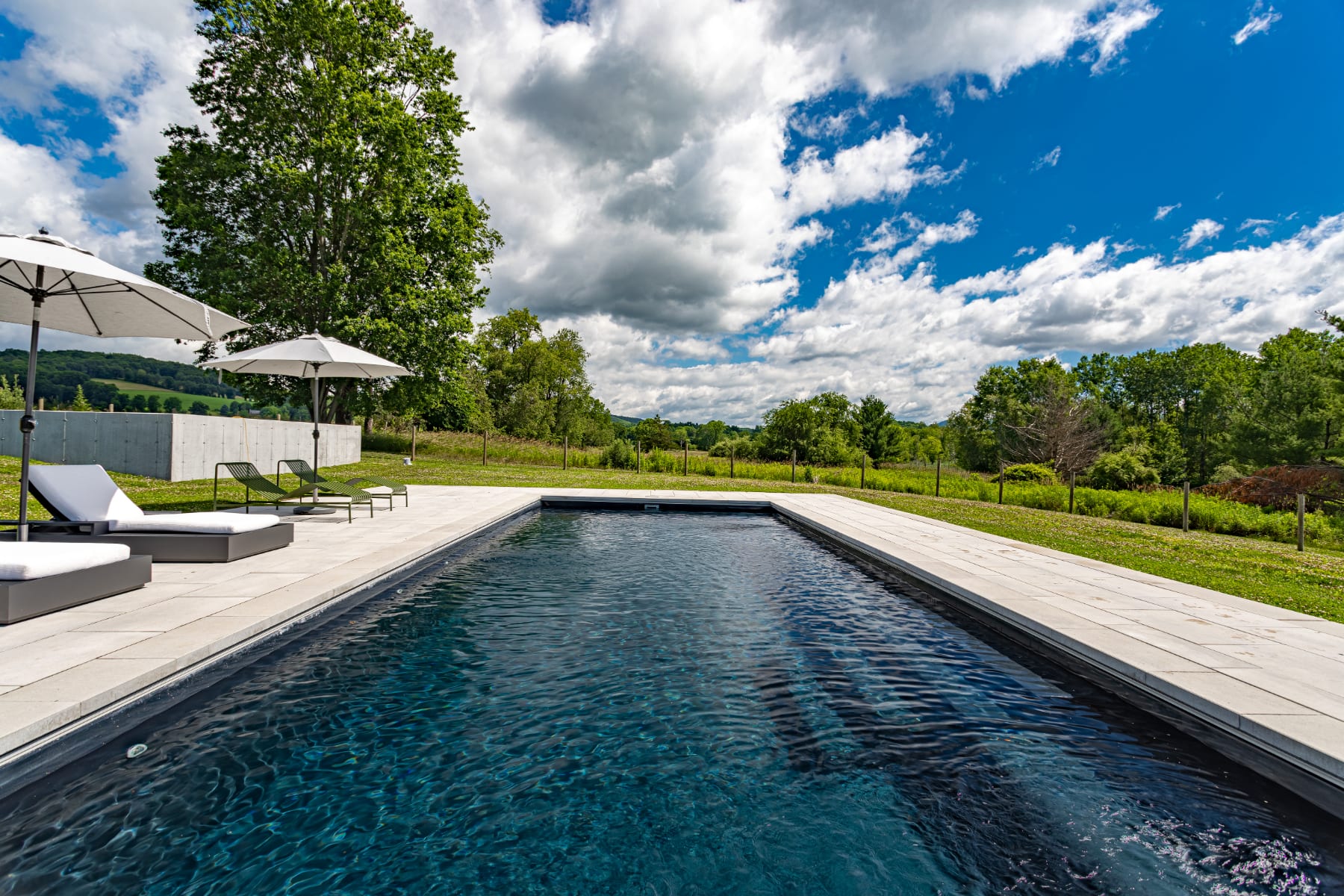 A long, dark-tiled swimming pool is bordered by light-colored pavers, with lounge chairs and umbrellas visible on the deck. The pool area is set against a backdrop of lush green hills, mature trees, and a bright blue sky with white clouds.