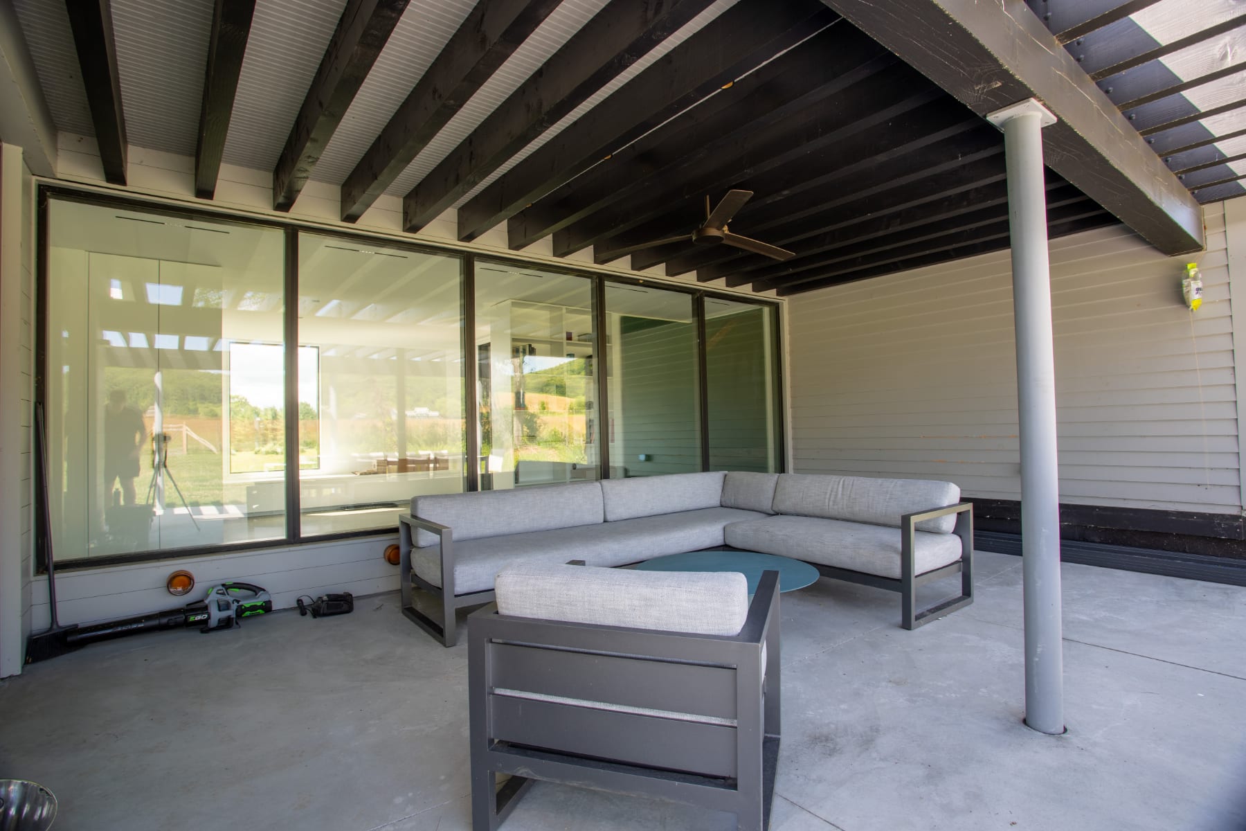 A contemporary outdoor patio features a large L-shaped sectional sofa and a single armchair with dark frames and light grey cushions, centered around a round blue coffee table. Large glass sliding doors connect to the interior, and a dark wooden pergola covers the space.