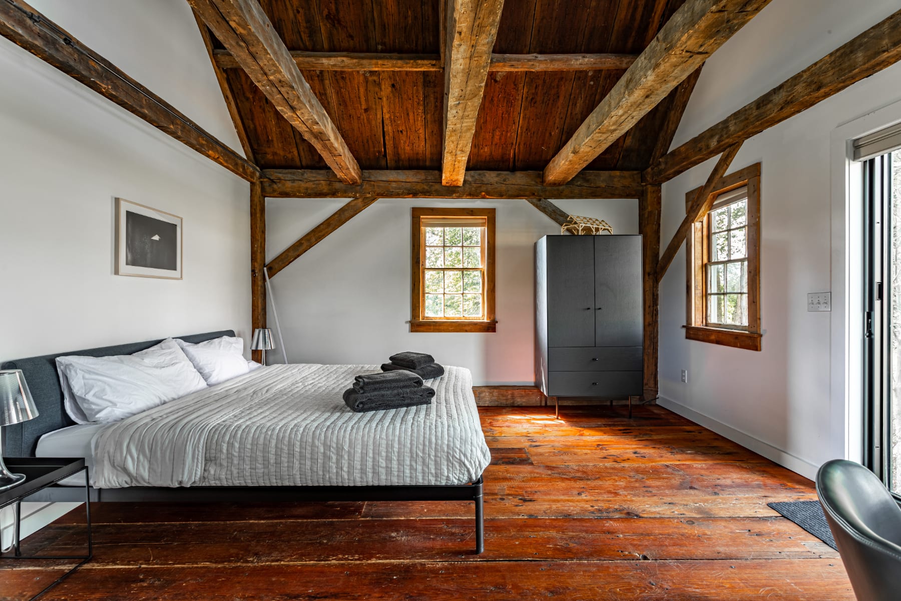 Cozy studio bedroom featuring a queen bed with white bedding and dark folded towels, a dark wardrobe, and rustic exposed wooden beams on the vaulted ceiling and walls.