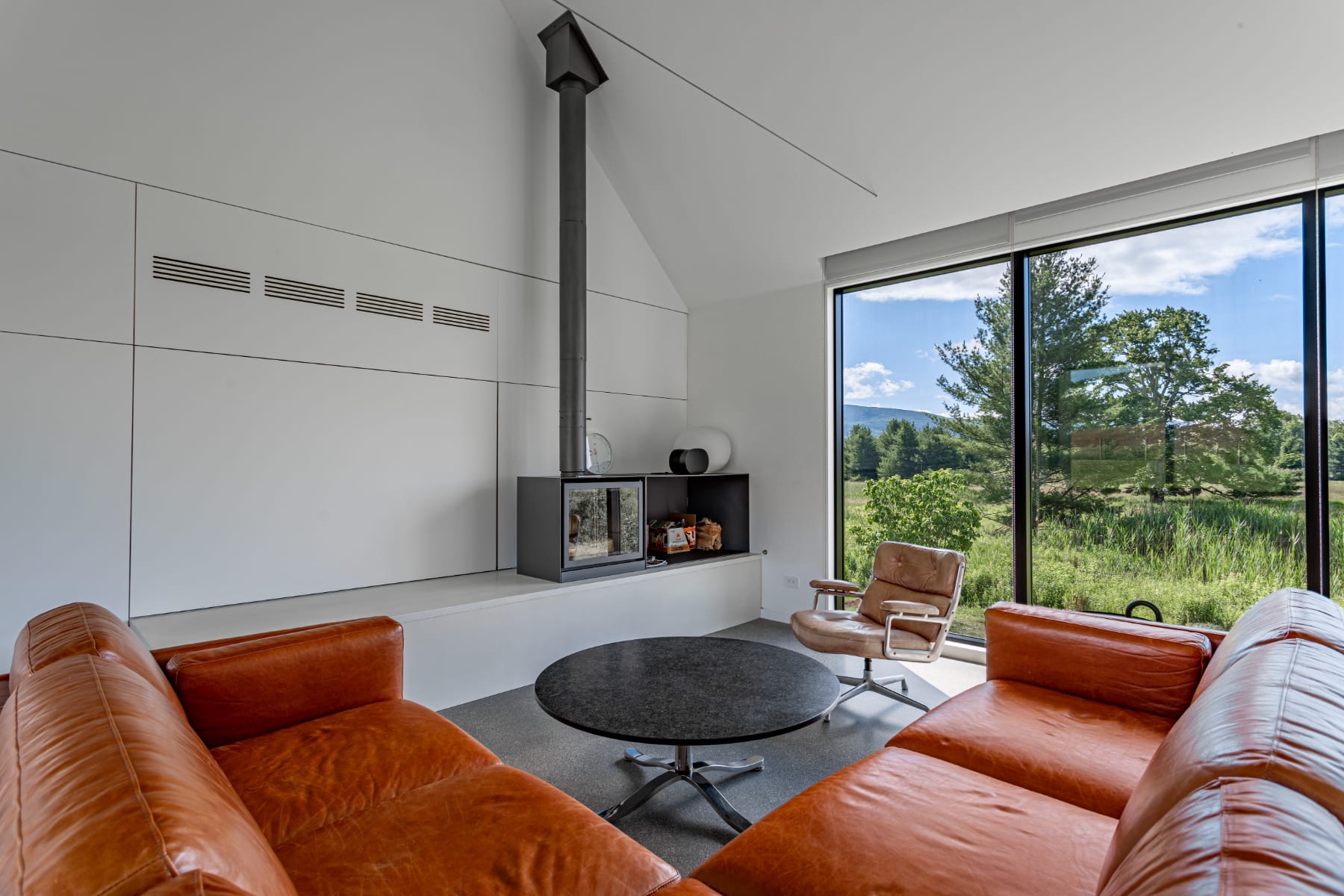 A contemporary living room features two large brown leather sofas arranged around a dark circular coffee table. A modern fireplace with a tall black chimney is integrated into white built-in shelving, with a large window offering scenic views of the outdoors.