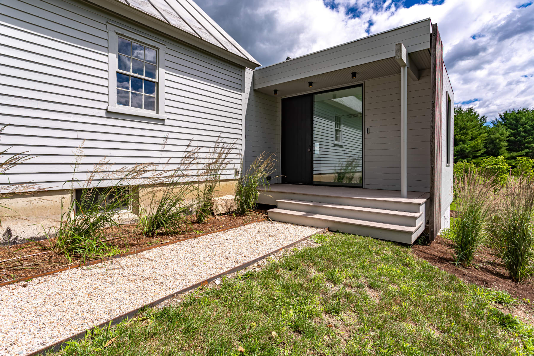 Exterior view of a light grey modern house with a dark entrance door and large glass panels, connected to an older, light-colored clapboard structure with a metal roof. A gravel path leads to the entrance, surrounded by grass and tall ornamental grasses.