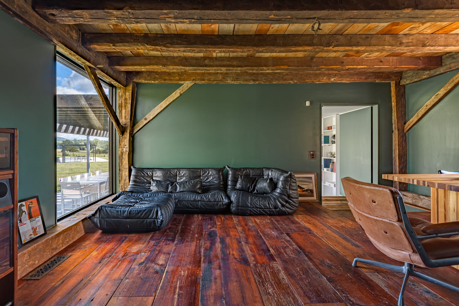 Rustic living area with a comfortable black leather sofa, dark green walls, and prominent exposed wooden beams on the ceiling and walls, complemented by rich hardwood floors and large windows.