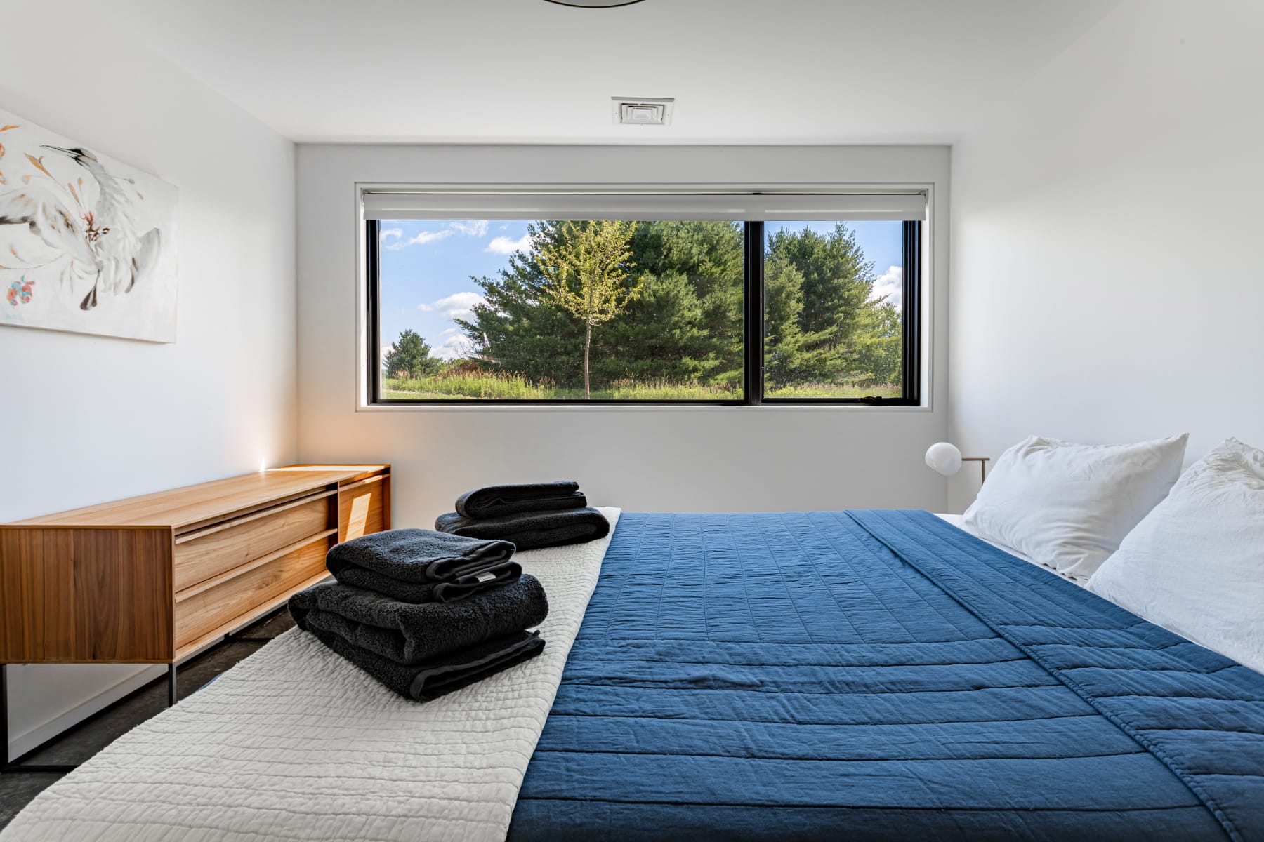 Another perspective of the bedroom, highlighting a sleek wooden dresser and a wide window framing a picturesque view of green trees and a bright sky. The bed is neatly made with dark blue and white linens.