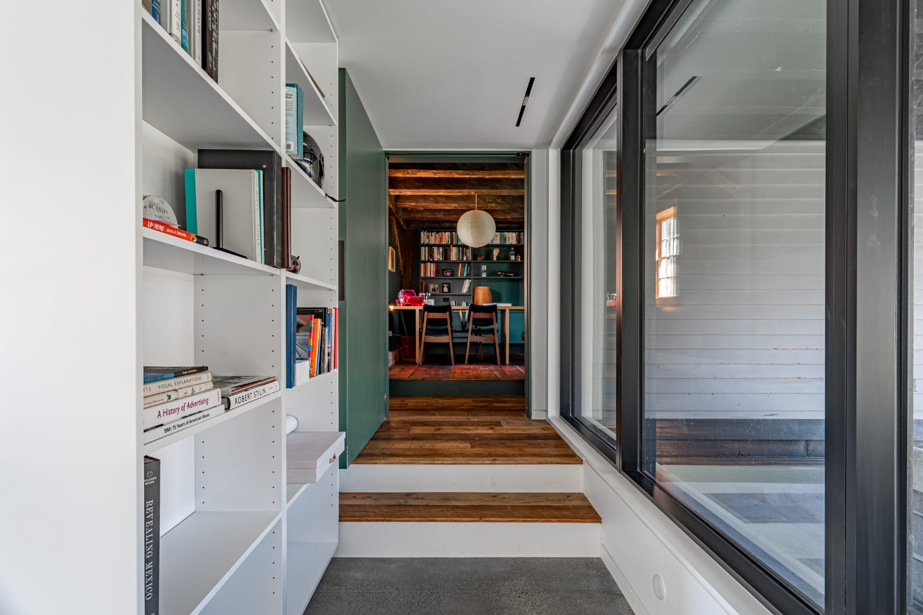 Bright hallway with white built-in bookshelves leading into a rustic office space, characterized by dark green walls, exposed wooden beams, and rich hardwood floors, with large windows providing natural light.