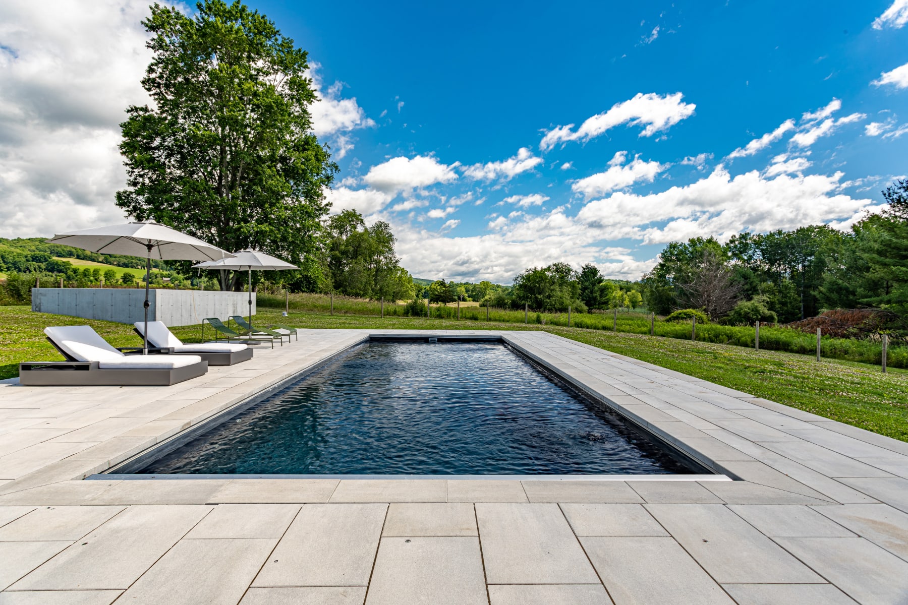 Modern rectangular swimming pool with dark water, surrounded by light grey pavers, set in a lush green landscape with distant hills under a blue sky. Two white lounge chairs with umbrellas are visible on the left.