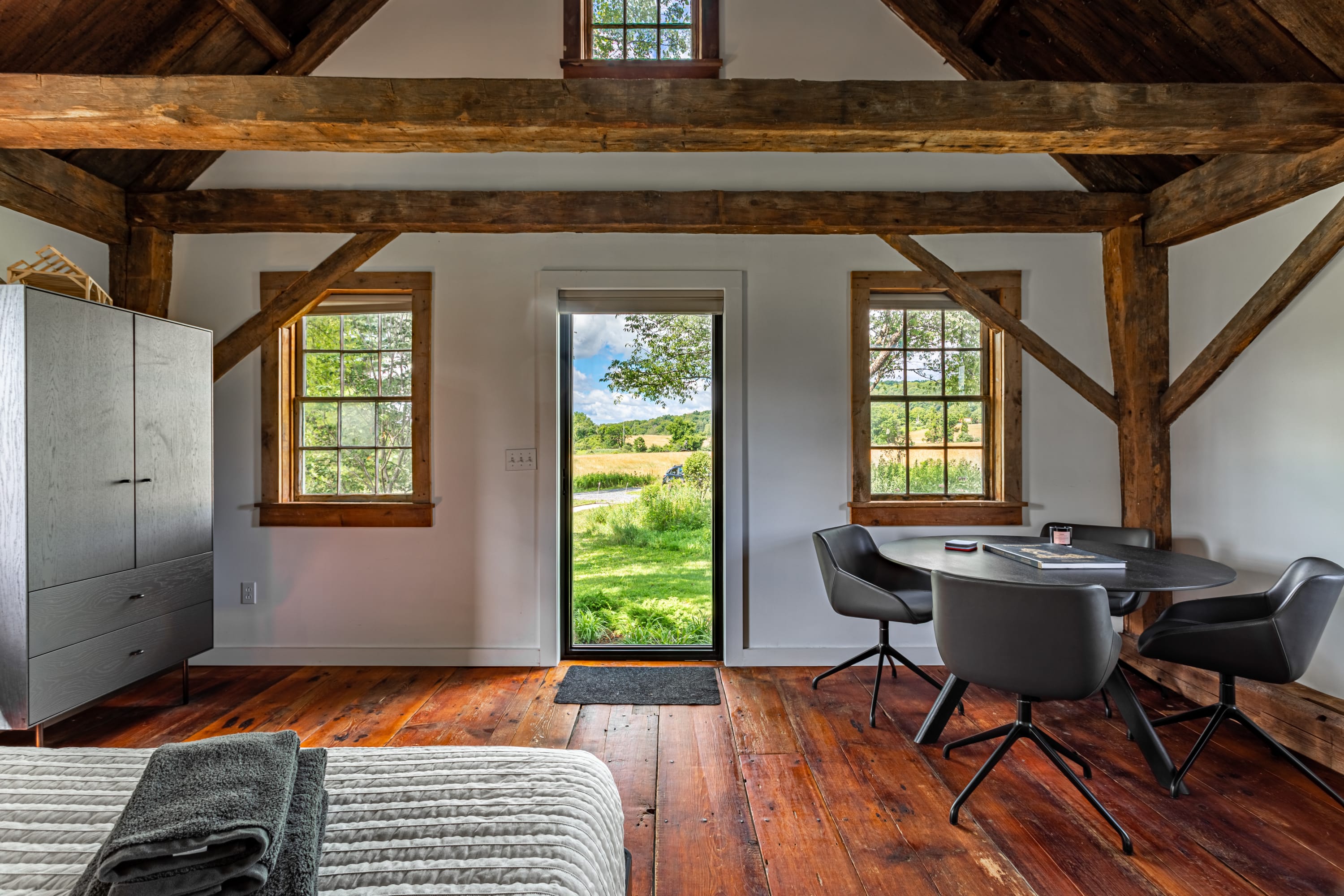 View of the studio bedroom showing the entrance door opening to a green landscape, a dark round dining table with chairs, and a dark wardrobe, all under exposed wooden beams.