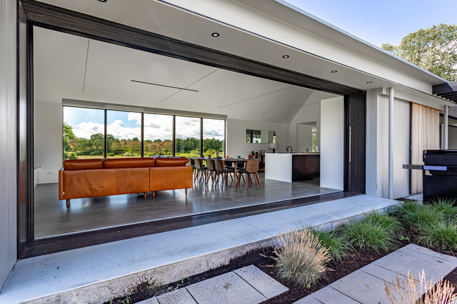 Expansive open-concept living and dining area with a large brown leather sofa, dark wood dining set, and polished concrete floors, opening via floor-to-ceiling glass doors to an outdoor patio and scenic landscape.
