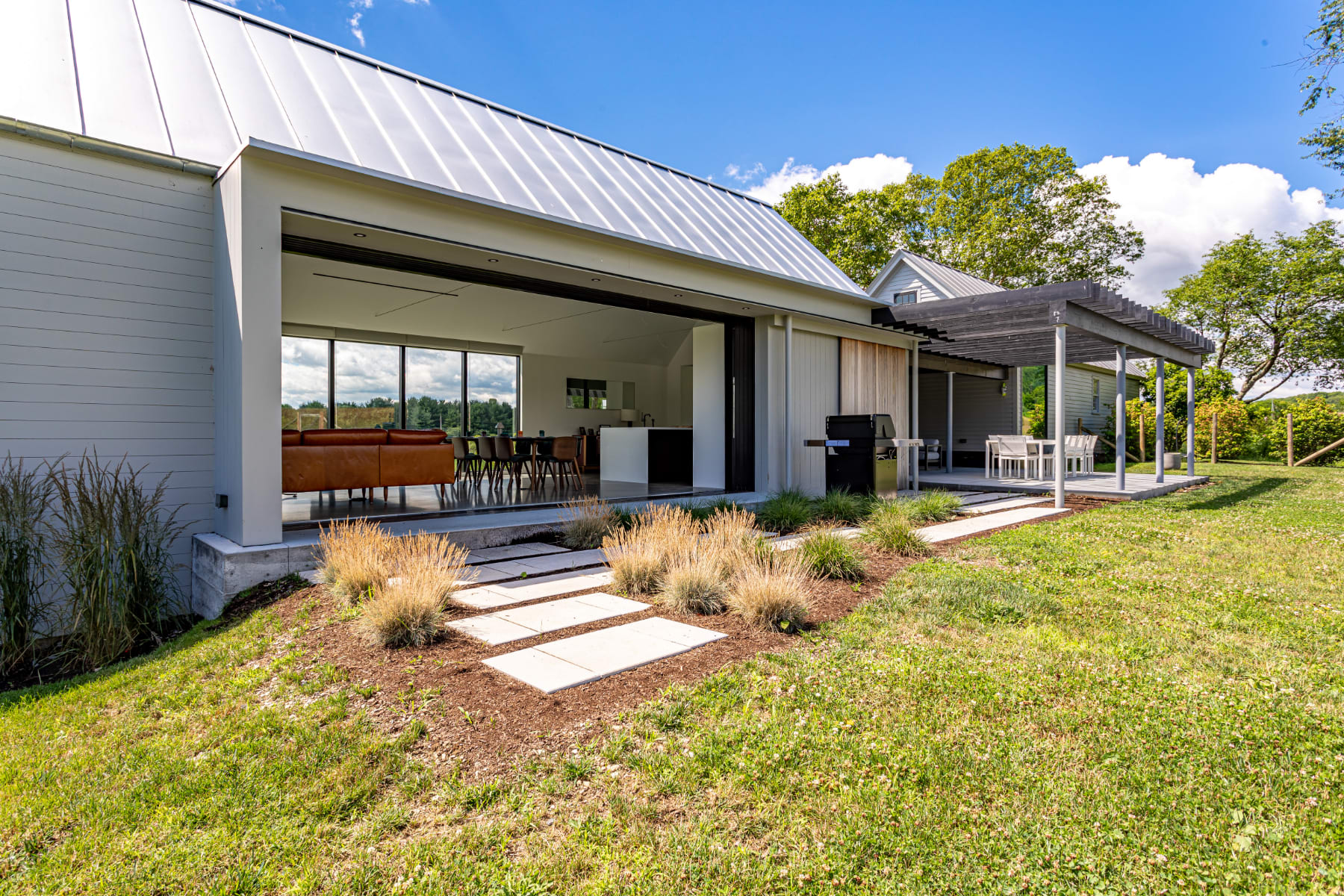 The modern house features a light-colored siding and a metal roof, with large glass sliding doors opening to a patio area. A BBQ grill and an outdoor dining set under a wooden pergola are visible, surrounded by landscaped grass and plants.