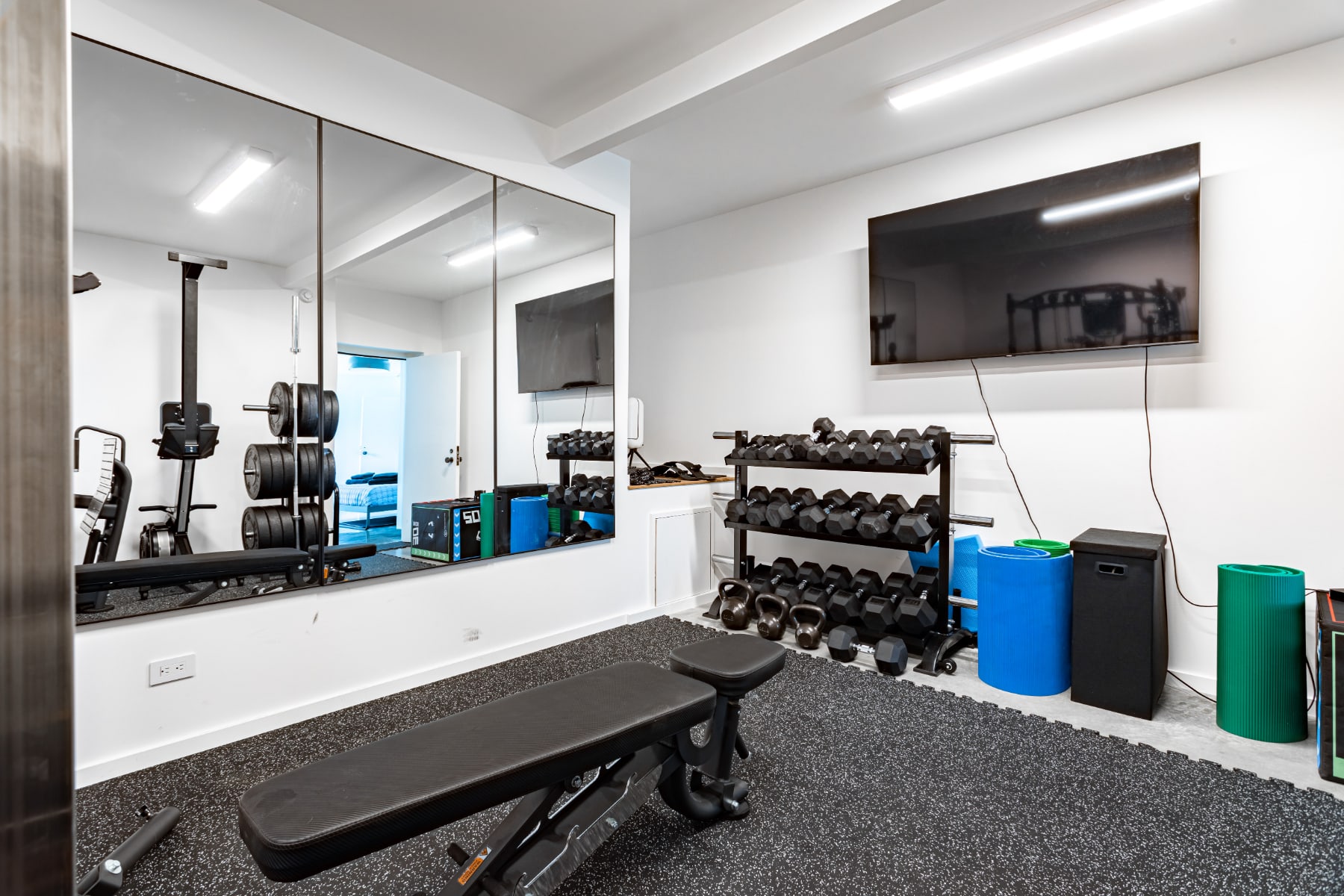 Spacious home gym equipped with a large mirrored wall, a rack of dumbbells and kettlebells, a weight bench, and various exercise mats. A large flat-screen TV is mounted on the white wall for entertainment.
