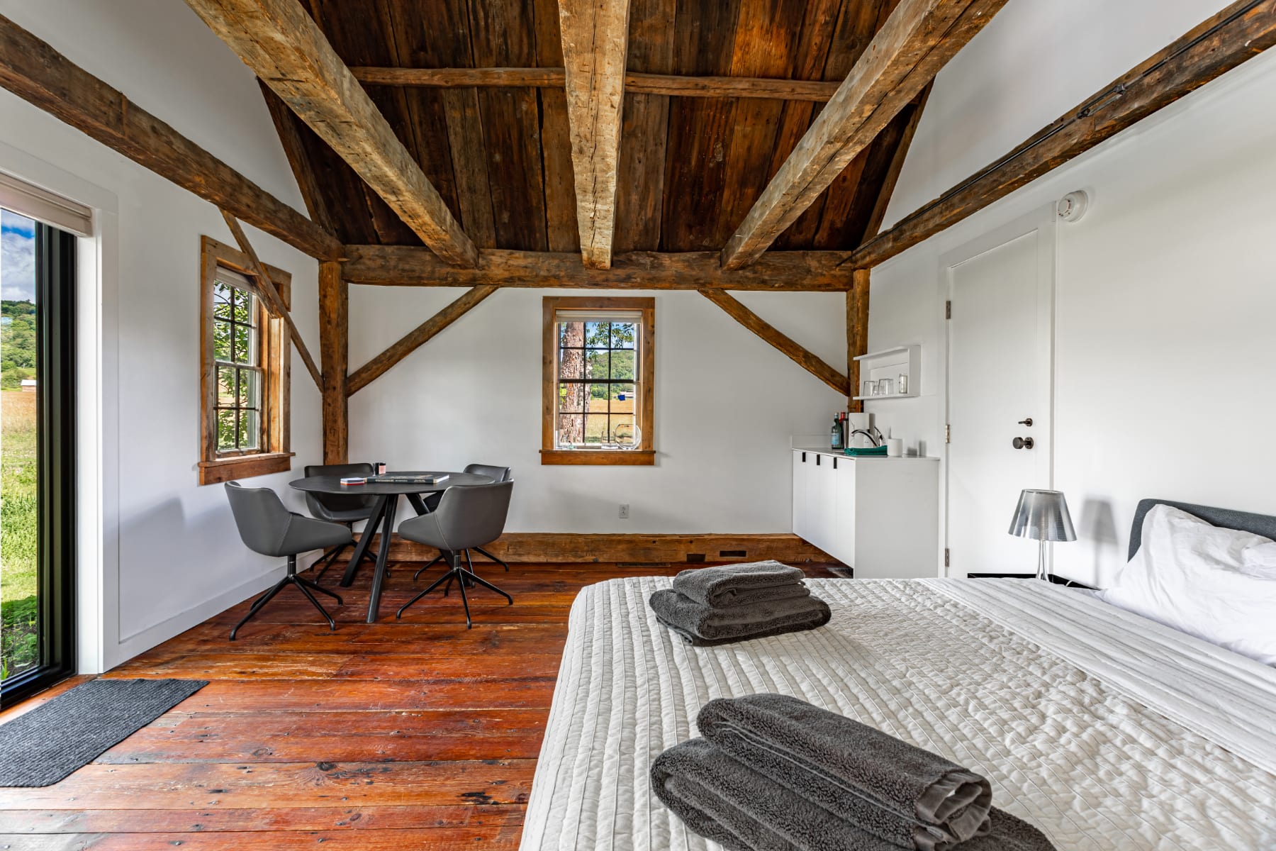 Bright studio bedroom with a queen bed, small dining table, and kitchenette, featuring rustic exposed wooden beams, vaulted ceiling, and rich hardwood floors.