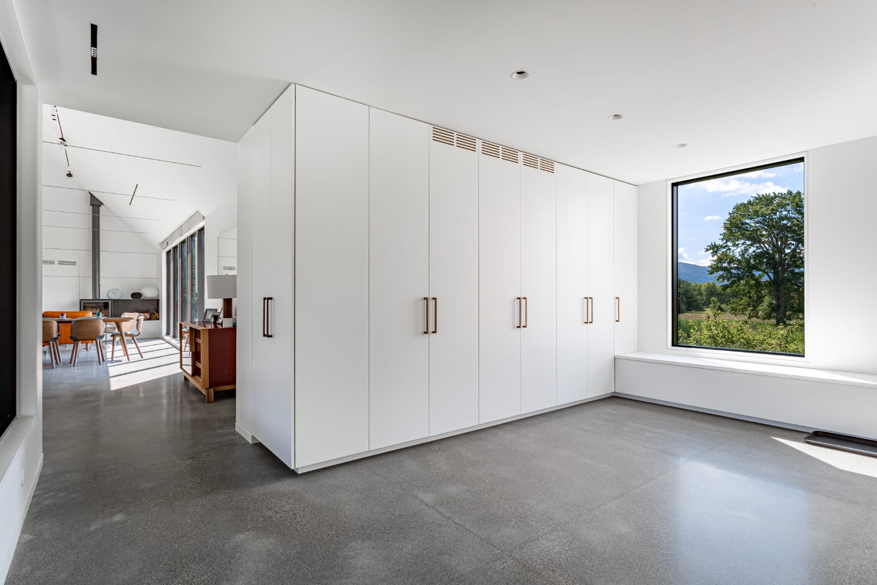Bright, minimalist room with polished concrete floors and a wall of white built-in cabinets with brass handles. A large floor-to-ceiling window offers a scenic view of a tree and green landscape, with a built-in bench below.