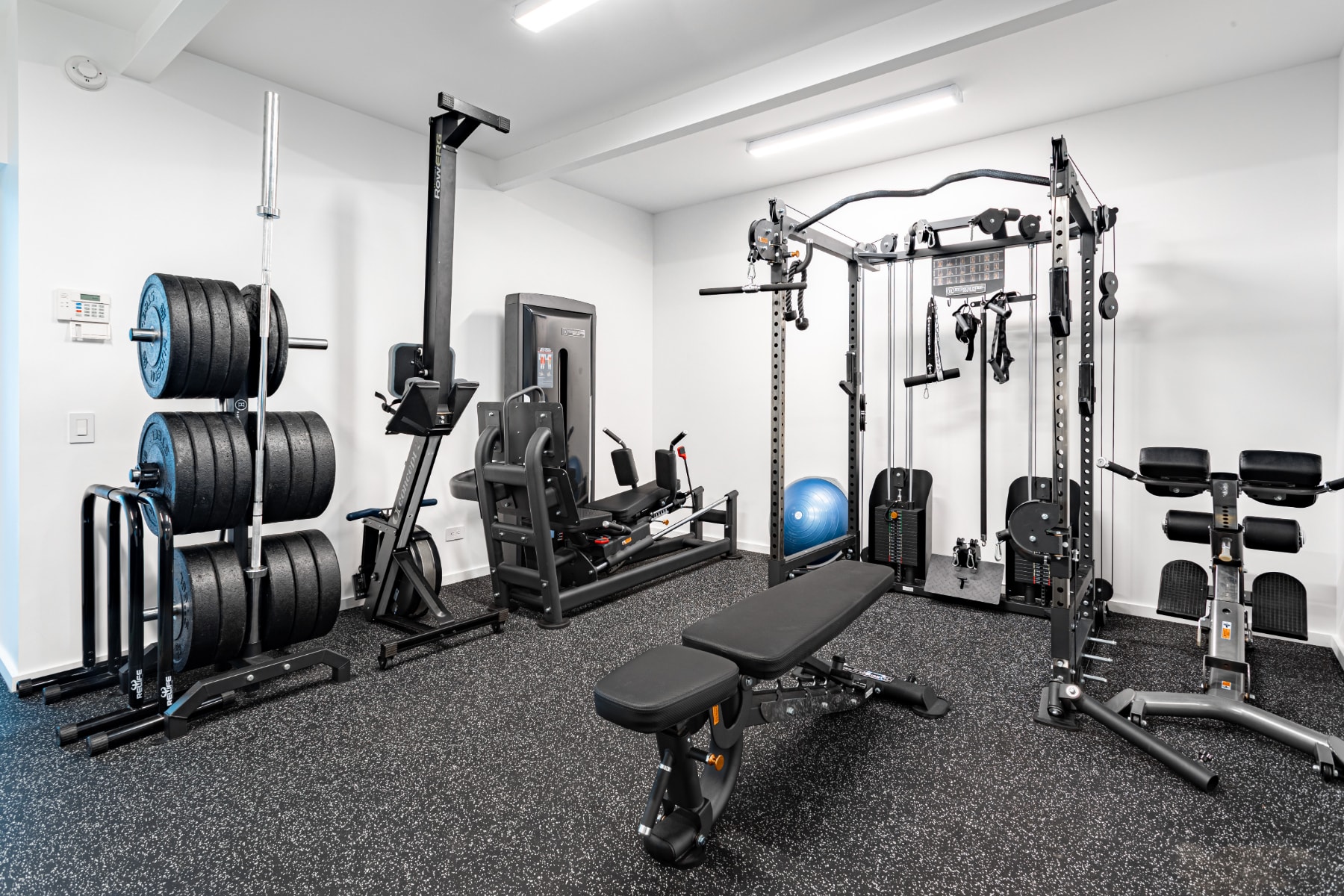 A well-equipped home gym features a variety of exercise machines including a weight rack with plates, a leg press, a multi-station gym, and a workout bench. The room has white walls and a black rubber floor.