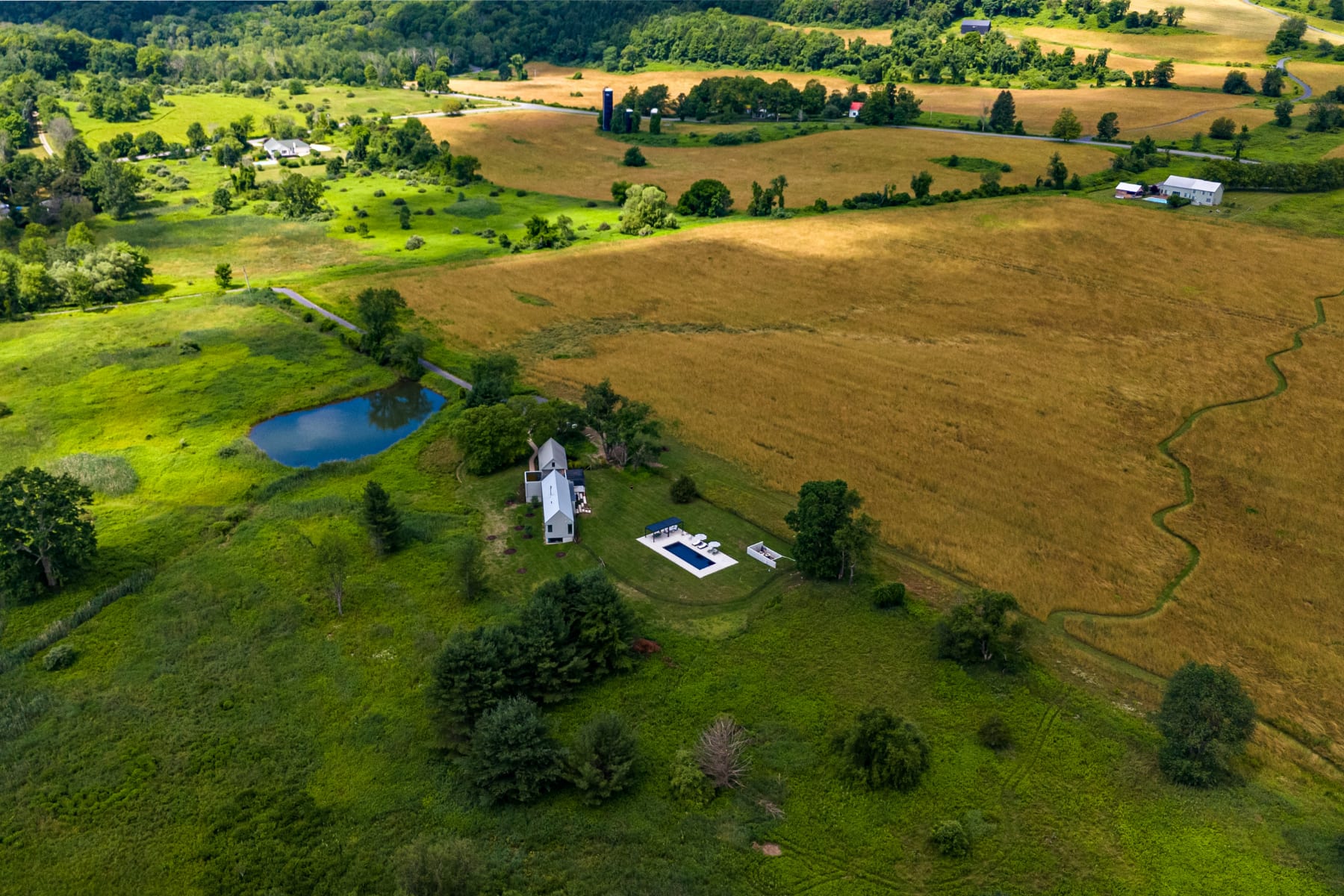 An aerial view showcases a modern house with a white roof, a rectangular swimming pool, and a small pond, nestled within expansive green fields and rolling hills under a clear sky. The property is surrounded by lush trees and distant farm structures.