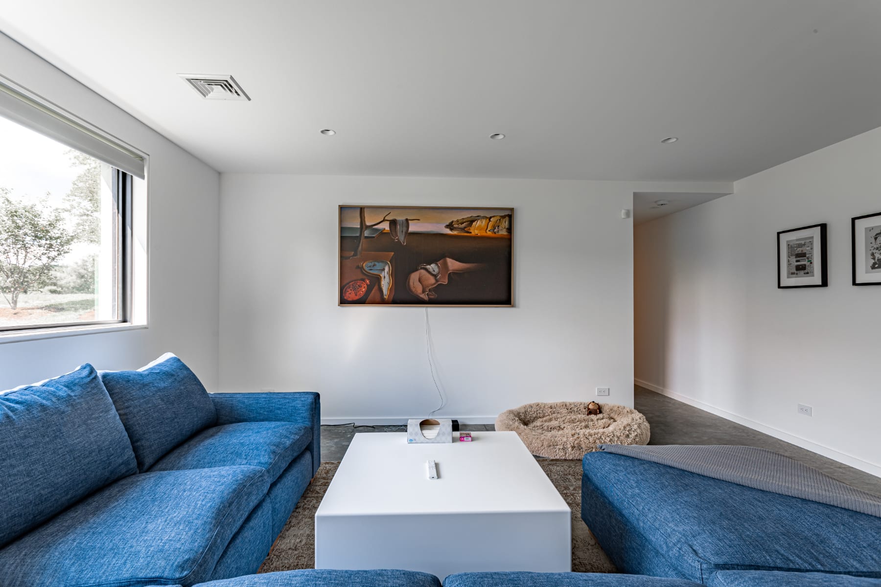 Bright living room with a blue sectional sofa, a white coffee table, and a distinctive artwork hanging on a white wall, with a pet bed visible on the concrete floor.