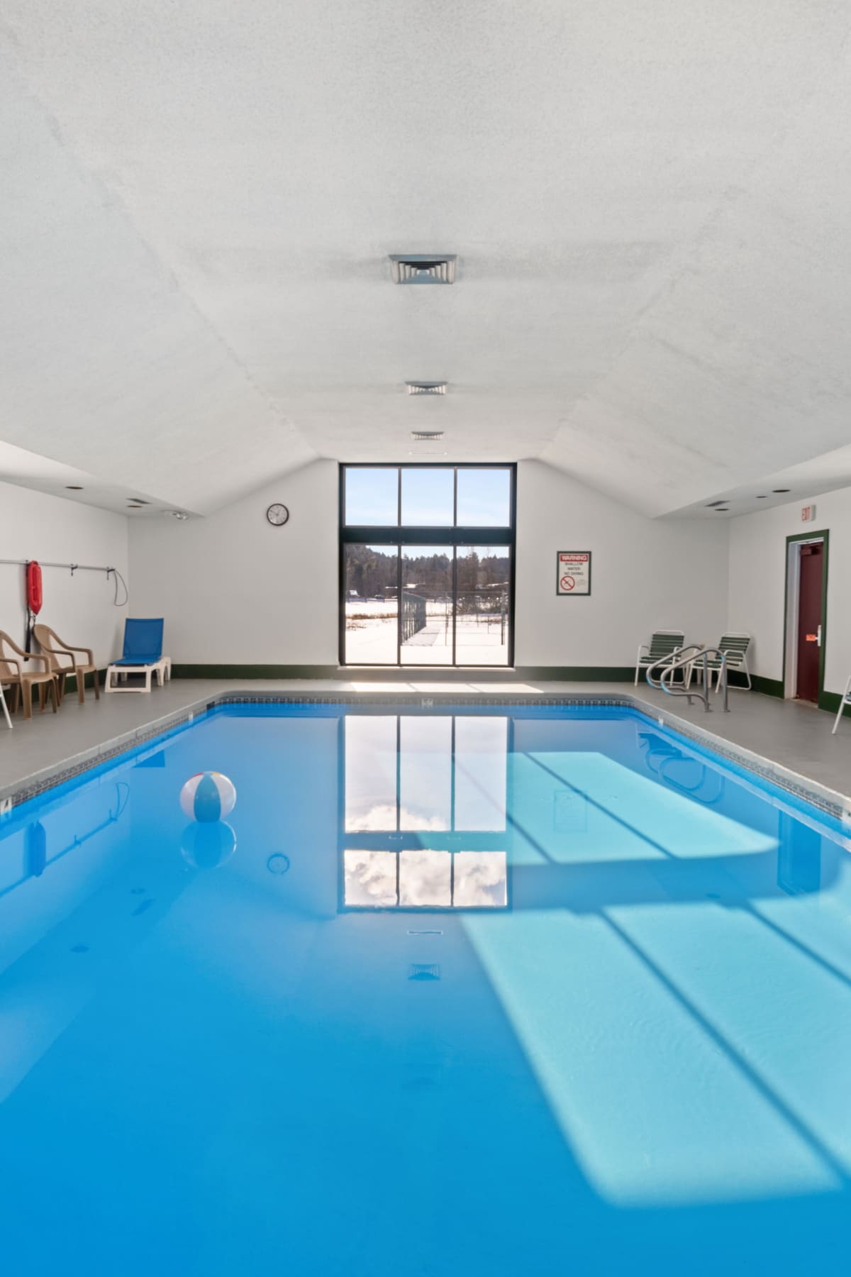 A bright indoor swimming pool with clear blue water and a white beach ball, reflecting the large window at the far end. Lounge chairs line the perimeter, and the ceiling is white with recessed lighting.