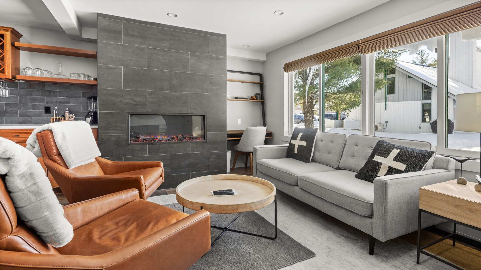 Cozy living room with a gray sofa, two brown leather armchairs, and a round wooden coffee table on a striped rug. A modern fireplace with a gray tile surround is a focal point.