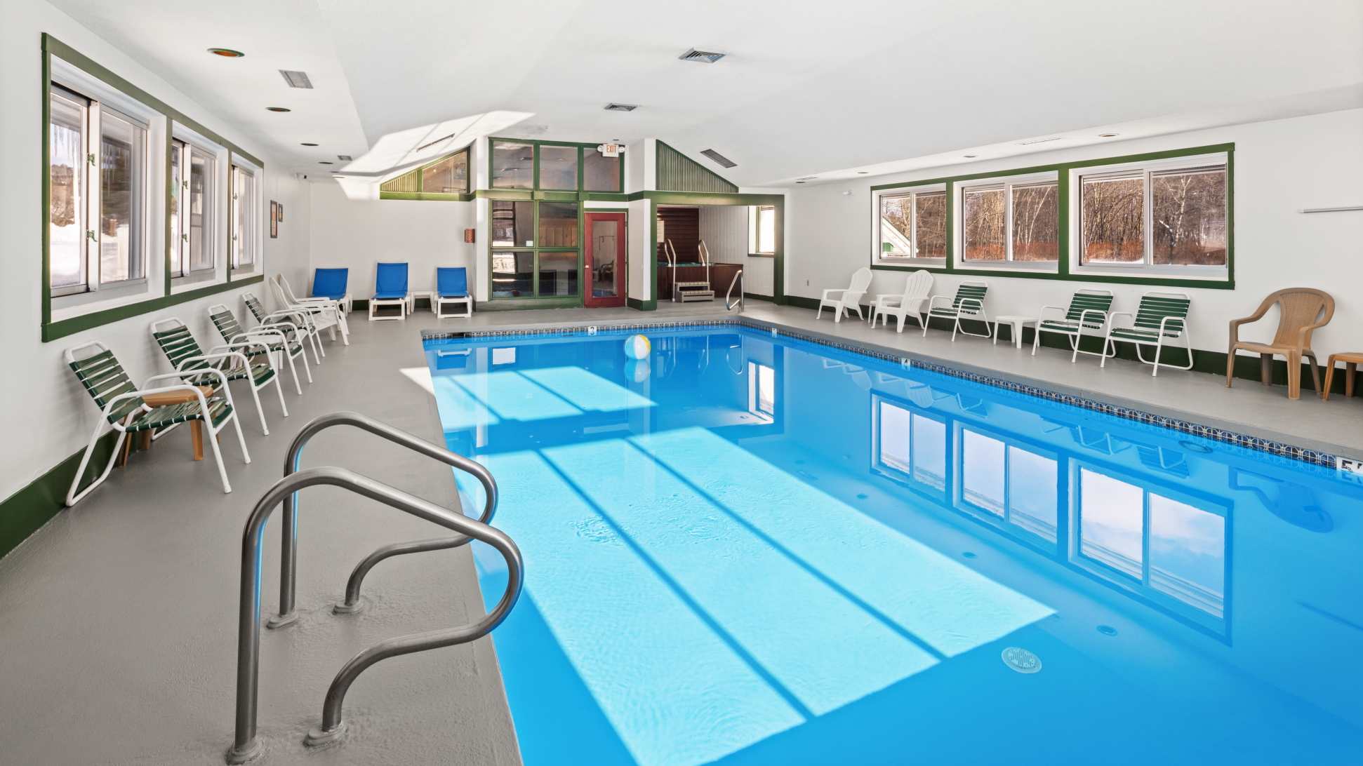 Bright indoor swimming pool area with a rectangular blue pool, surrounded by lounge chairs and large windows looking out onto a snowy landscape. The room features white walls and green trim around the windows and ceiling.