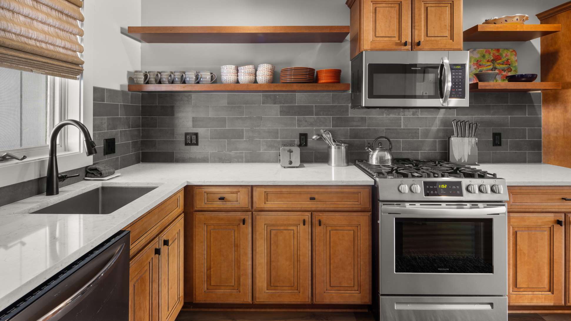 Corner view of a modern kitchen featuring light wood cabinets, white countertops, and a gray subway tile backsplash. Stainless steel appliances include a gas range, microwave, and dishwasher.