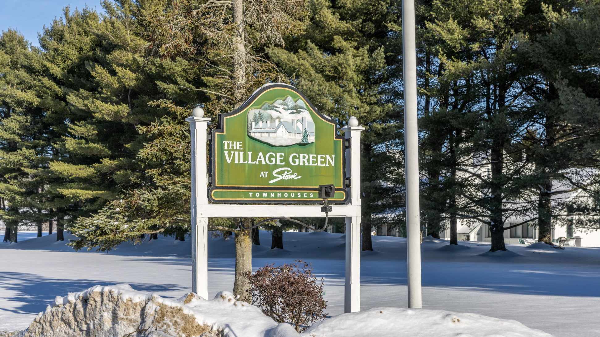 A prominent green and white arched sign for "The Village Green at Stowe Townhouses" stands on white posts in a snow-covered landscape. Tall evergreen trees frame the sign under a bright sky, indicating the property entrance.