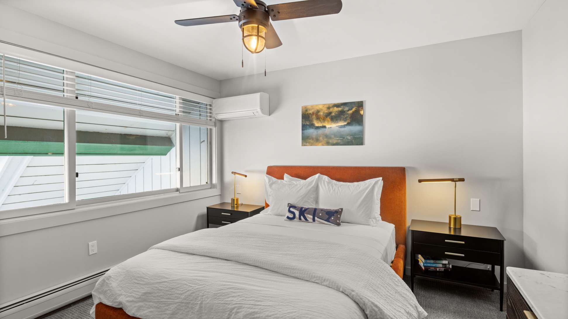 Cozy bedroom with an orange upholstered headboard, white bedding, and two black nightstands with gold-accented lamps. A large window with blinds and a ceiling fan are visible.