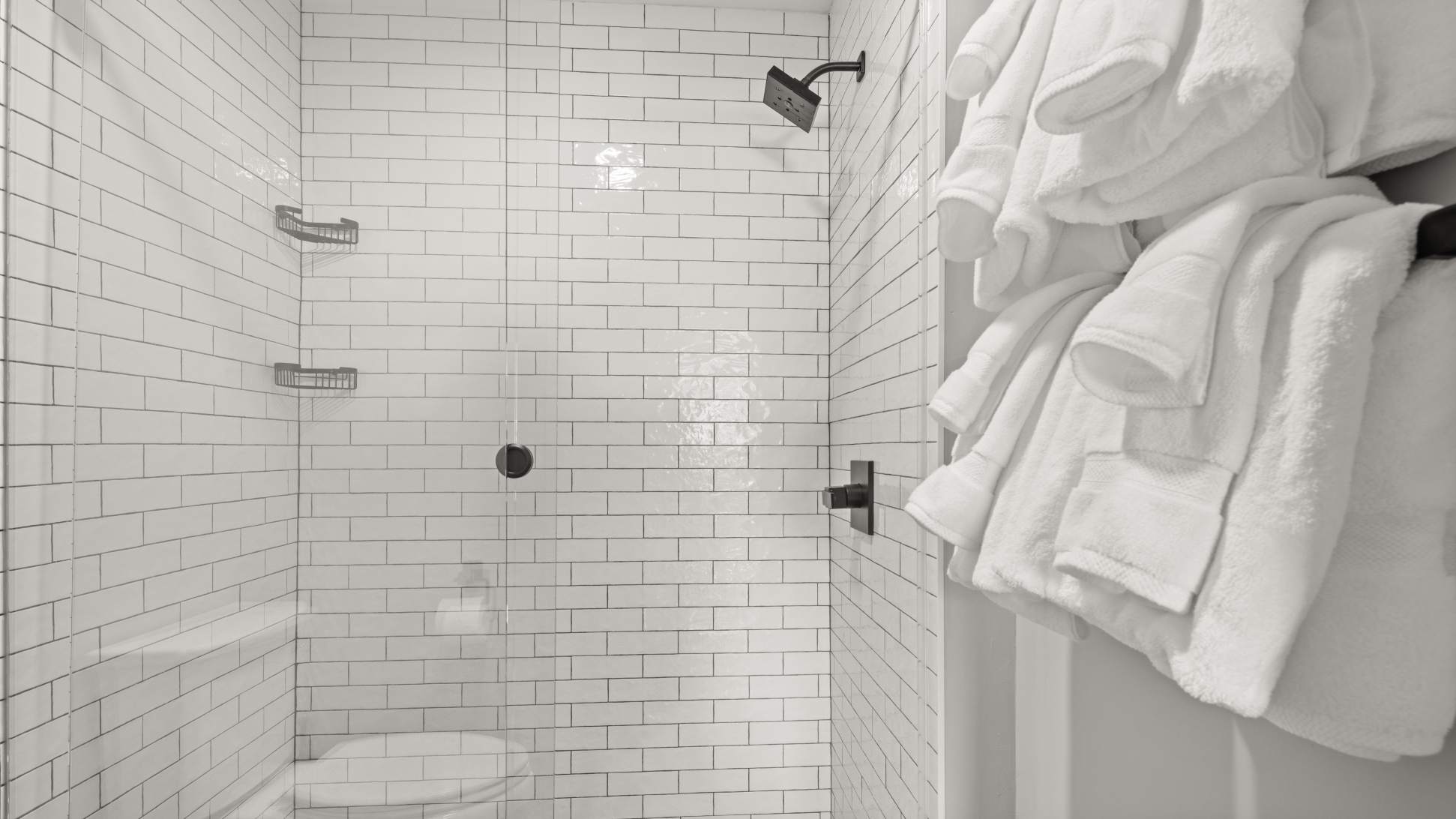 A clean, modern shower with white subway tiles, a black shower head, and a niche for toiletries. Fresh white towels are neatly folded and hung nearby.