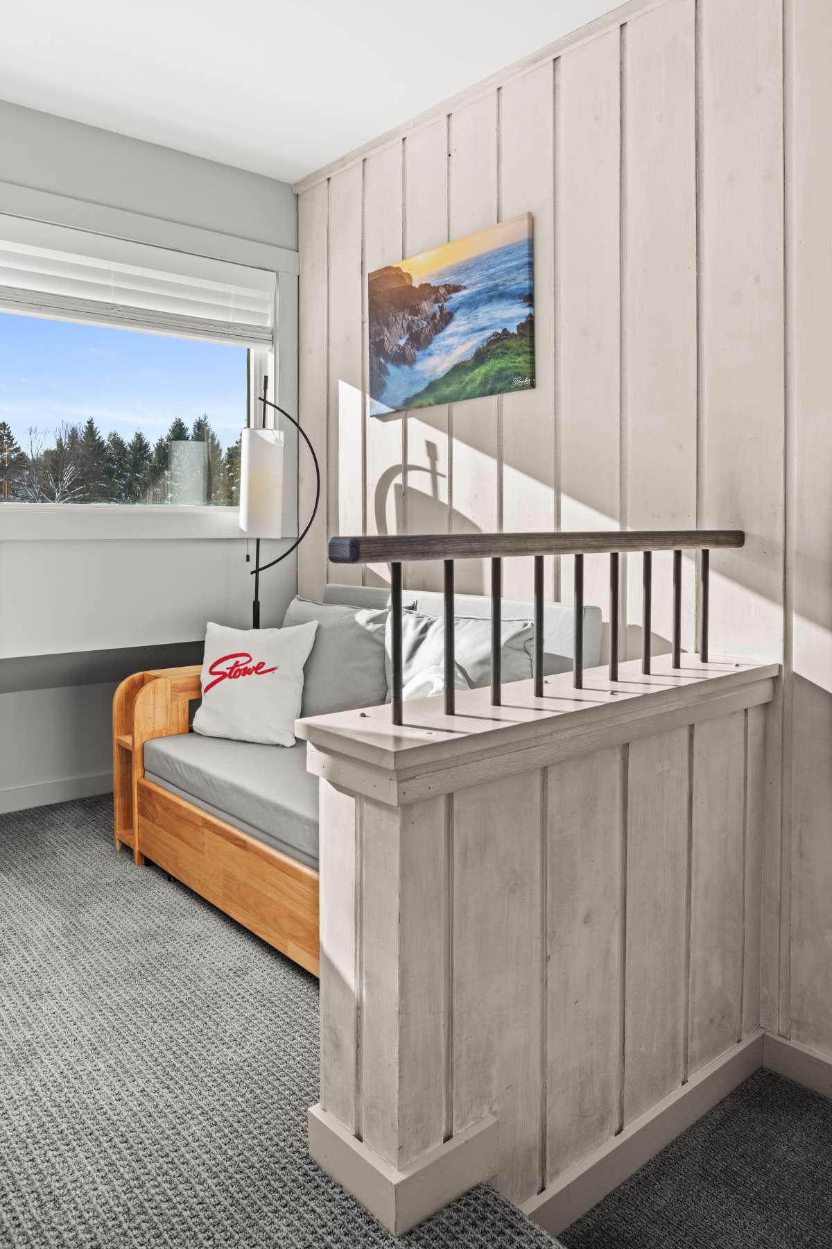 A cozy sitting area features a wooden daybed with gray cushions and a white 'Stone' pillow, set against a light wood-paneled wall. A large window offers views of a snowy landscape, and a modern floor lamp provides light.