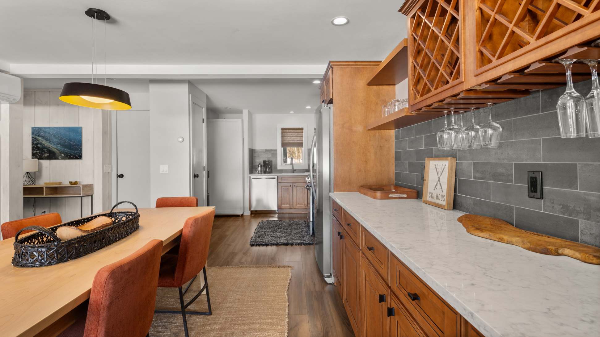 View from the dining area towards the kitchen, showing a light wood dining table with orange chairs. The kitchen features light wood cabinets, stainless steel appliances, and a wet bar with a wine rack.