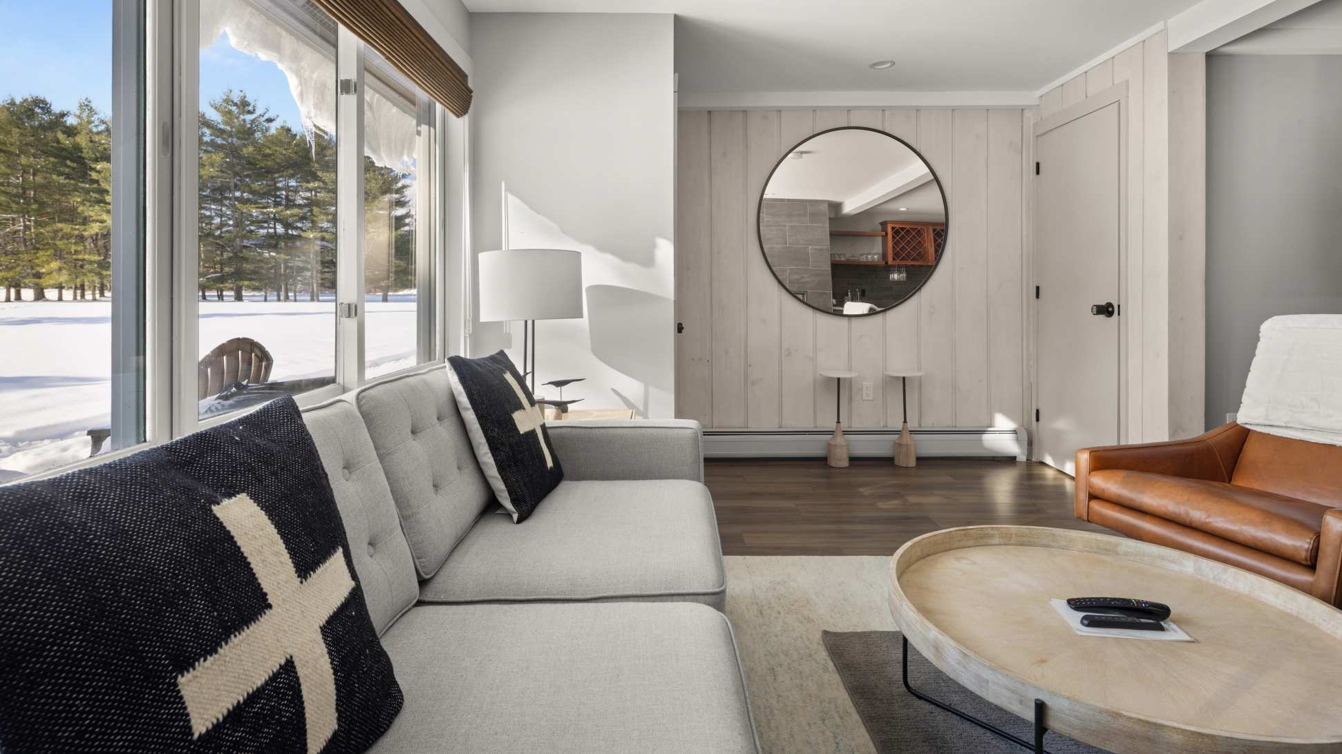Bright living room with a gray sofa, a round wooden coffee table, and a large window overlooking a snowy landscape. A circular mirror reflects the adjacent kitchen area.