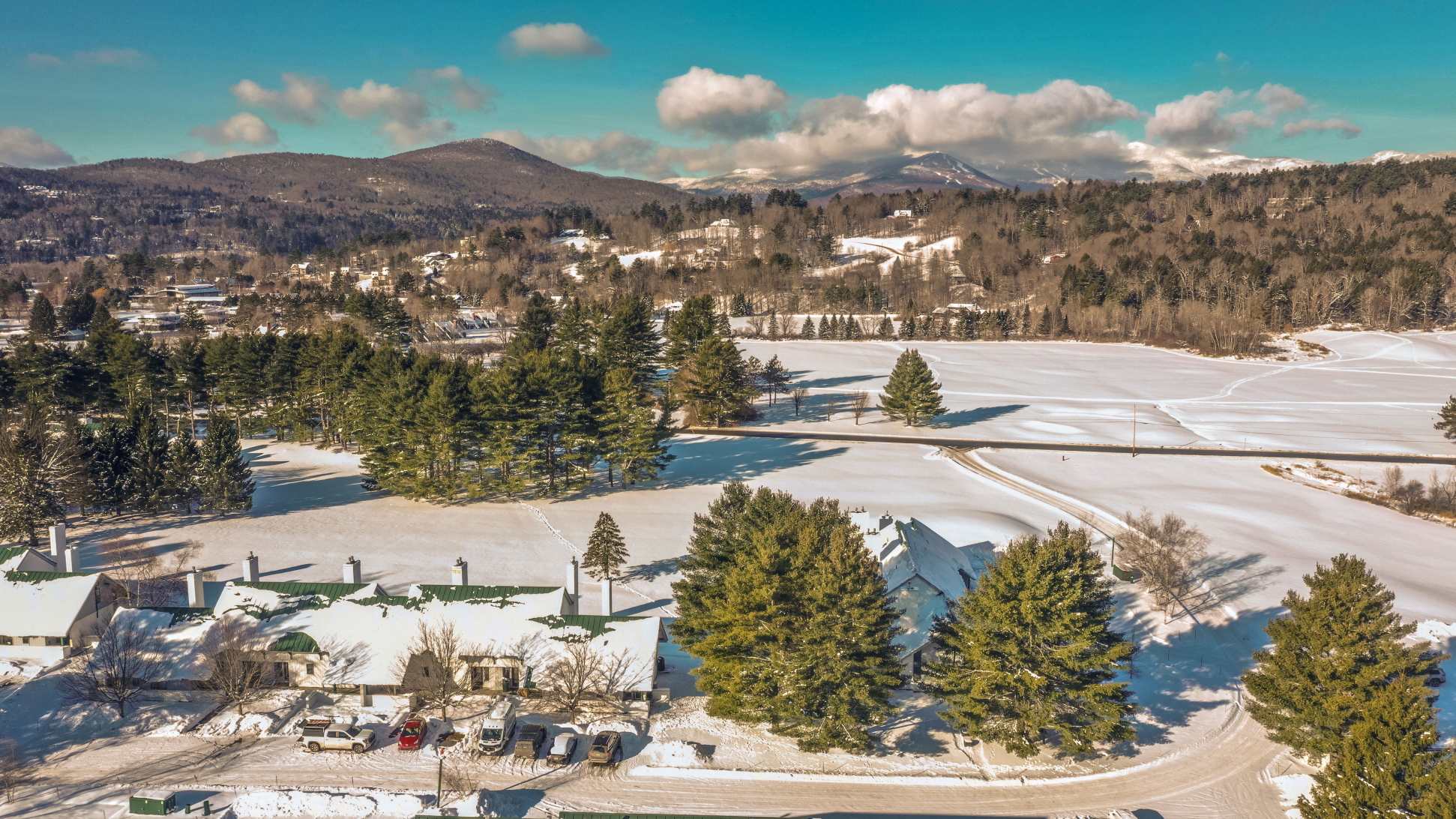 An aerial view captures a cluster of buildings with distinctive green roofs, surrounded by vast snow-covered grounds and tall evergreen trees, with a road winding through the landscape. Distant snow-capped mountains rise under a partly cloudy blue sky, showcasing a serene winter vista.