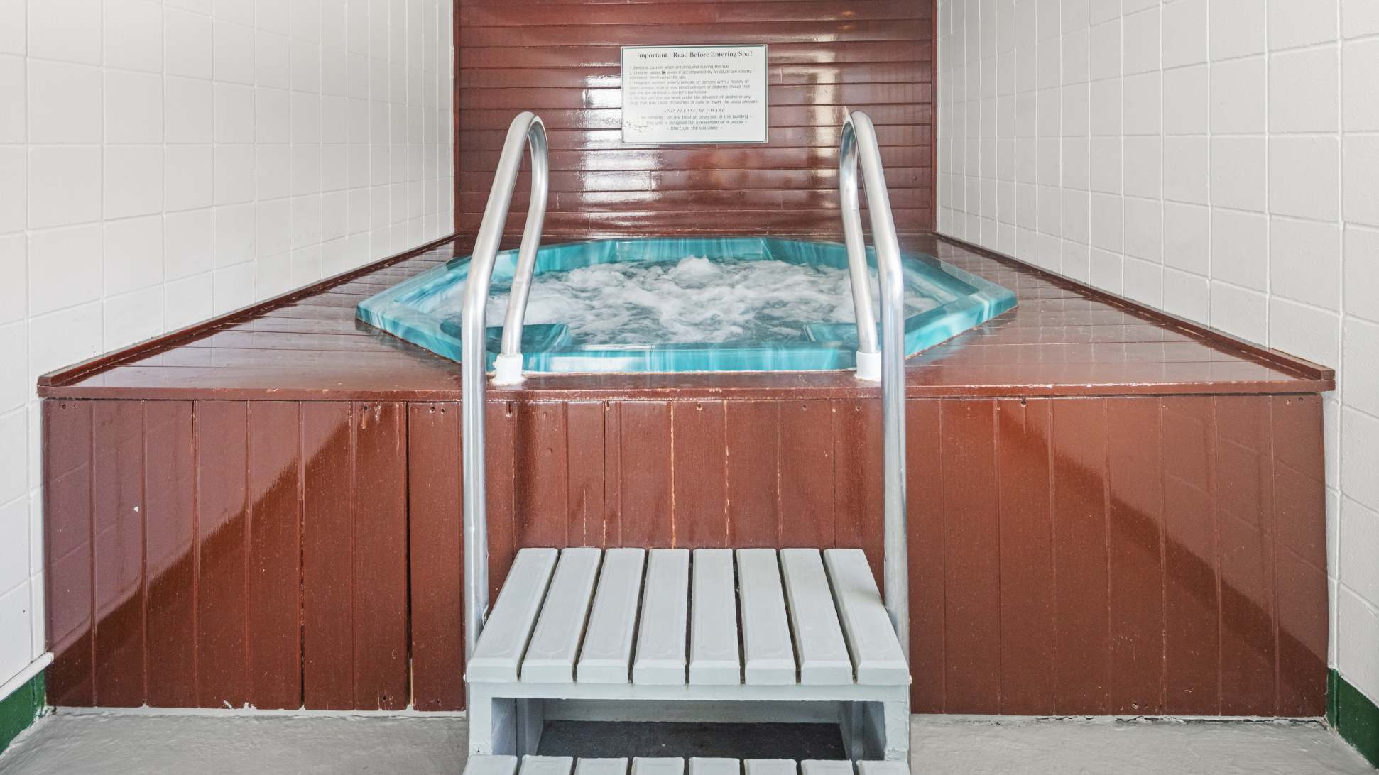 An indoor hot tub is set into a dark wood surround, with white tiled walls and metal handrails for entry. The water is bubbling, indicating it's in use.