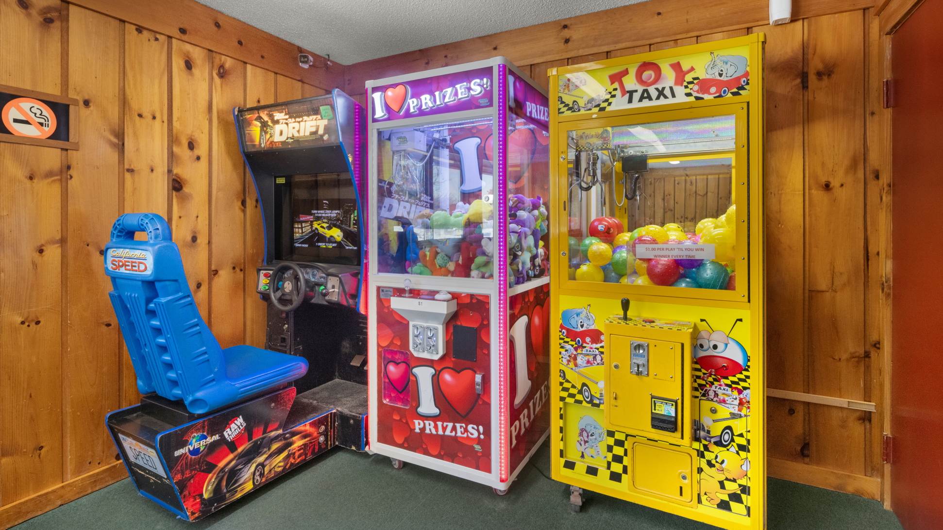 A corner of a game room features three arcade machines: a racing game, a "I Love Prizes" claw machine, and a "Toy Taxi" claw machine, all set against wood-paneled walls.