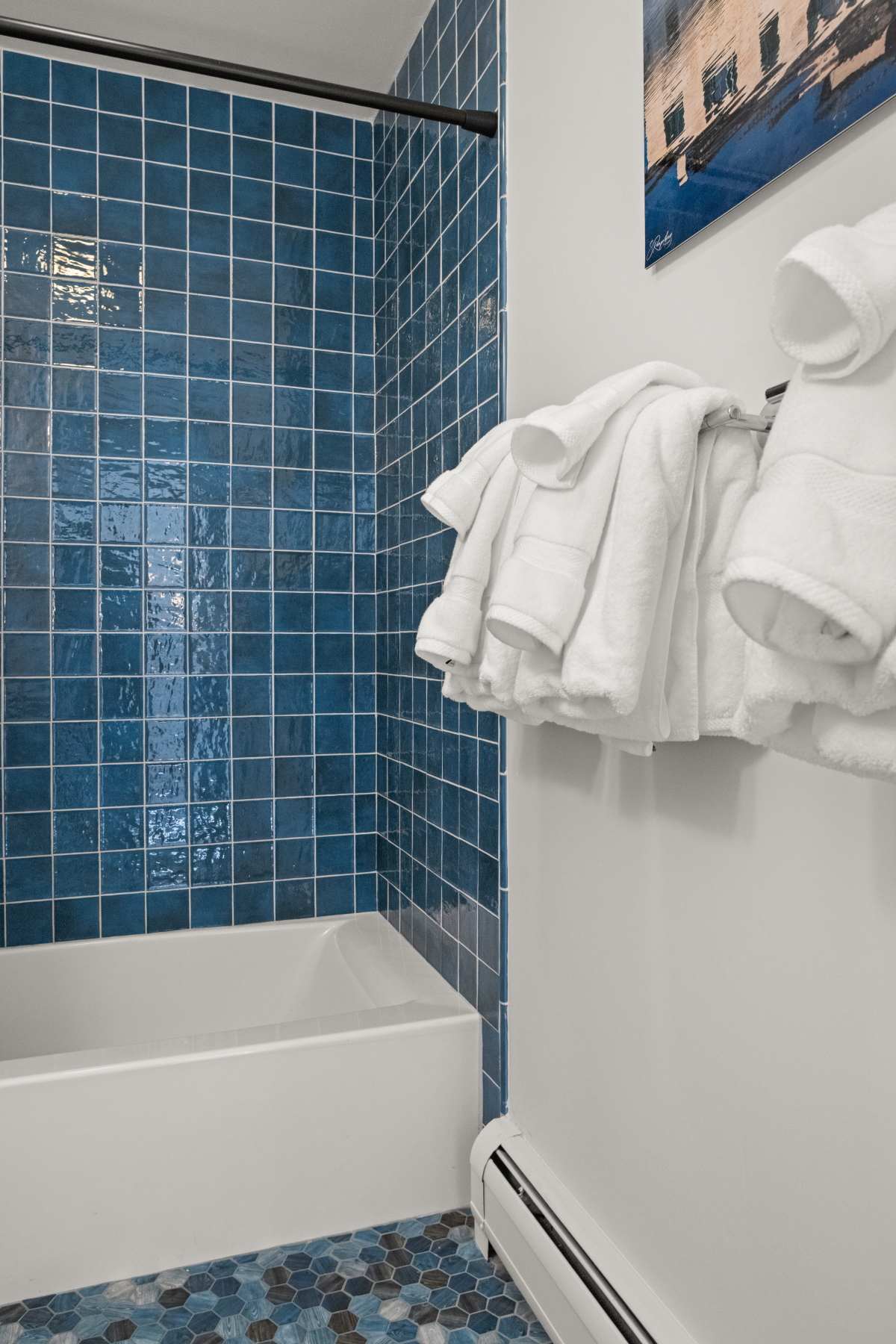 Close-up of a bathroom's shower/tub combo, featuring vibrant blue square tiles and a contrasting blue hexagonal floor tile, with white towels hanging on a rack.