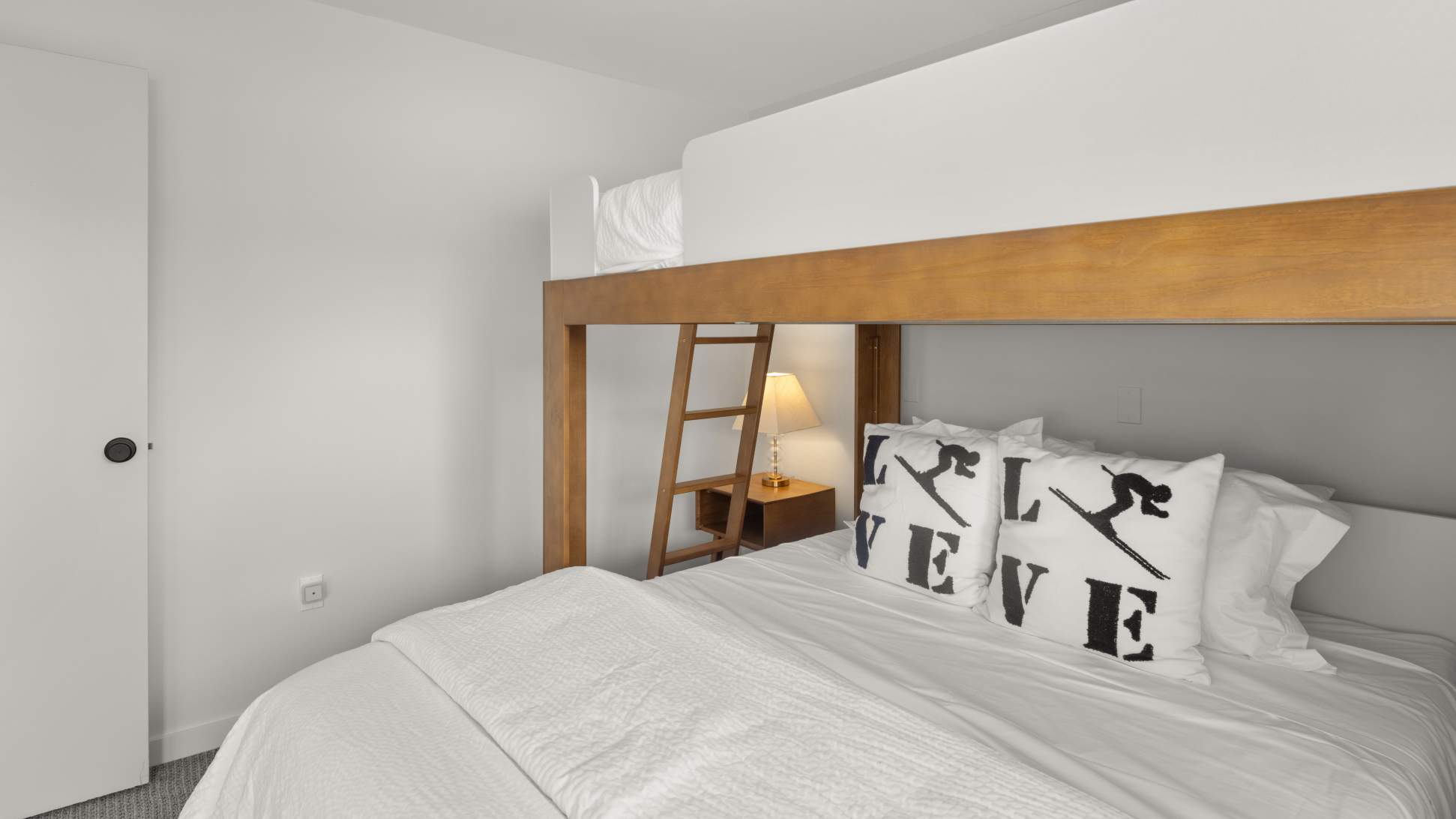 A bright bedroom featuring a wooden bunk bed frame with a queen-sized bed on the bottom and a twin bed on top, adorned with white bedding and "Stowe" pillows.