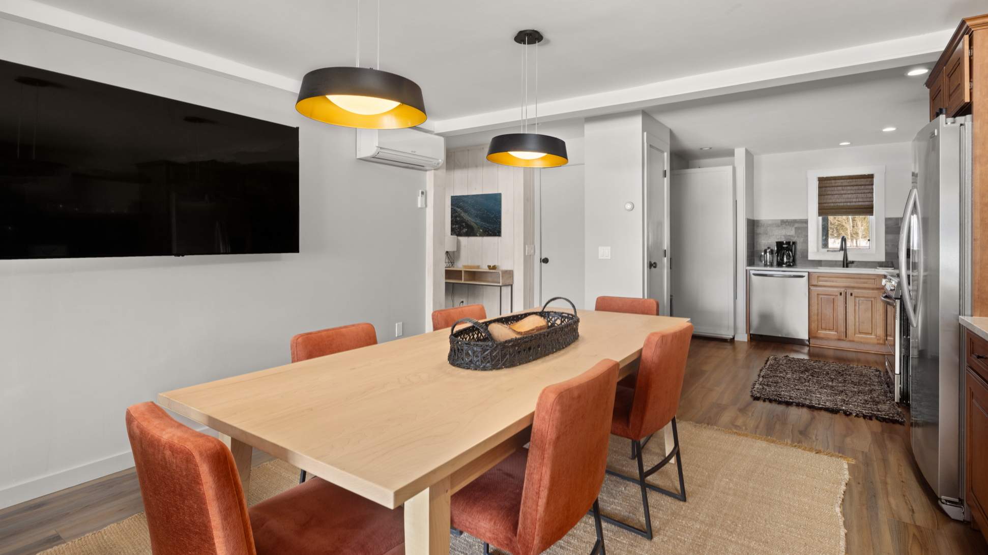 Bright dining area with a light wood table, six orange upholstered chairs, and two modern black and gold pendant lights. A large wall-mounted TV is visible on the left.