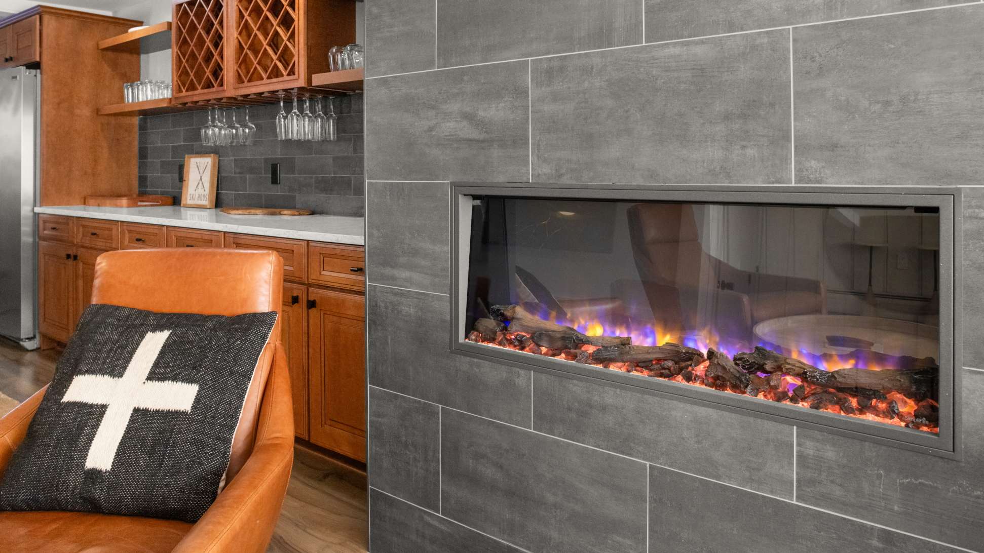 Close-up of a modern electric fireplace set into a gray tiled wall, displaying colorful artificial flames and logs. A brown leather armchair with a decorative pillow is visible to the left, next to a wooden wet bar area.