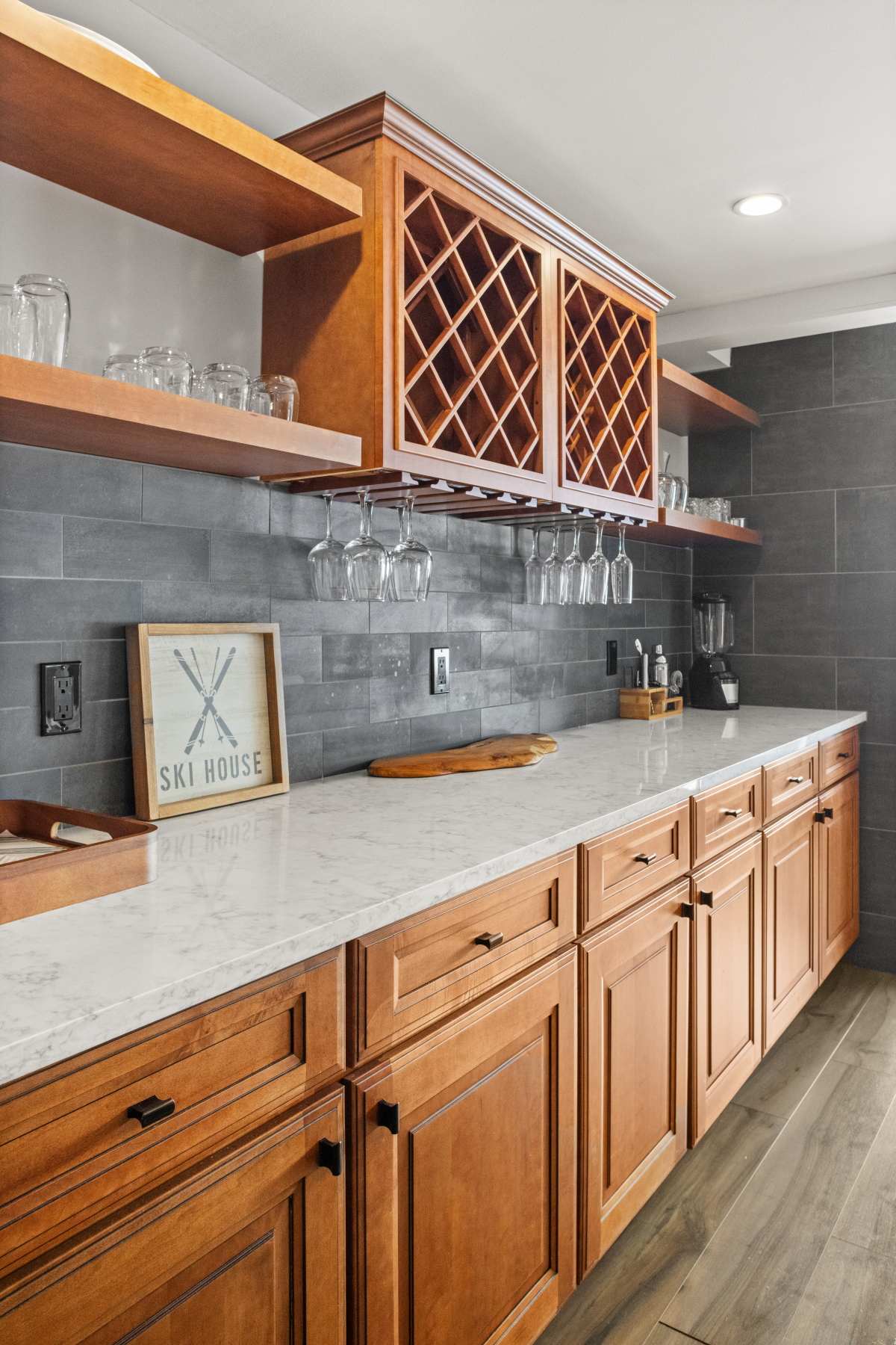 A stylish wet bar area features light wood cabinetry, a white marble countertop, and gray subway tile backsplash. Open shelving holds glassware, and a built-in wine rack with hanging wine glasses adds to the sophisticated design.