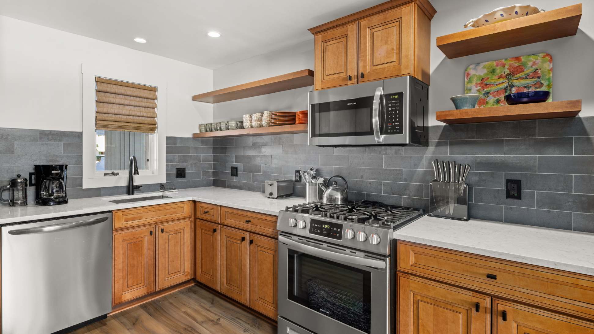 Modern kitchen with light wood cabinetry, white quartz countertops, a gray subway tile backsplash, and stainless steel appliances including a gas range and microwave. Open shelving displays dishes.