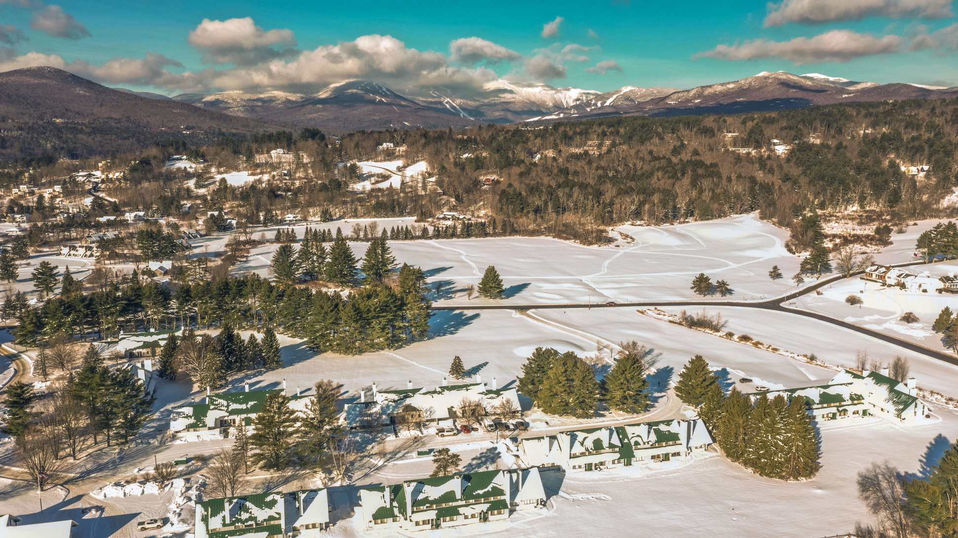 A wide aerial shot reveals a sprawling complex of green-roofed buildings nestled among snow-covered fields and evergreen forests, with a road cutting through the winter landscape. Majestic snow-capped mountains form a dramatic backdrop under a bright blue sky with scattered clouds.