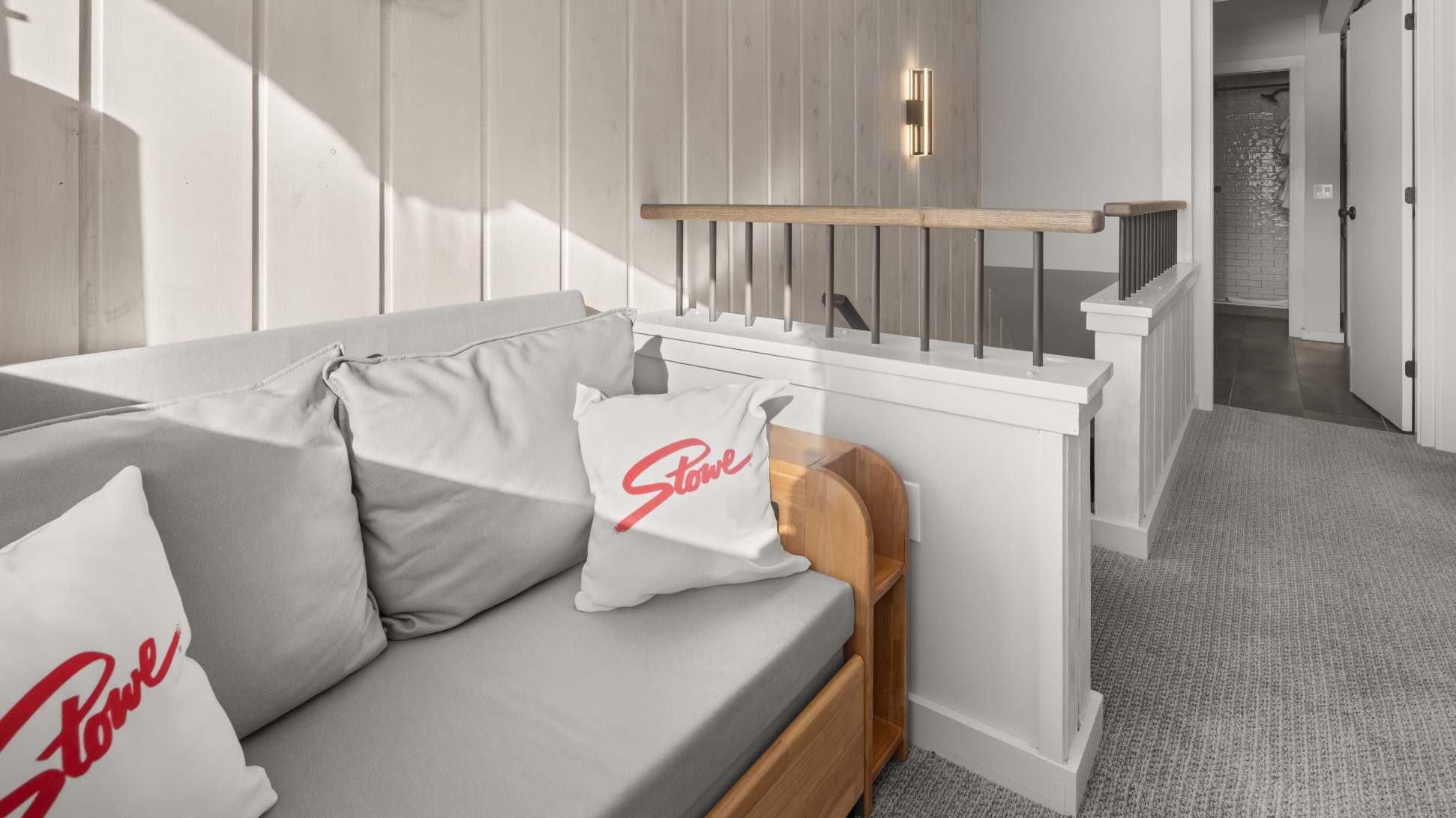A bright loft area features a light gray daybed with "Stowe" branded pillows, set against white vertical wood paneling, next to a modern railing overlooking a staircase.