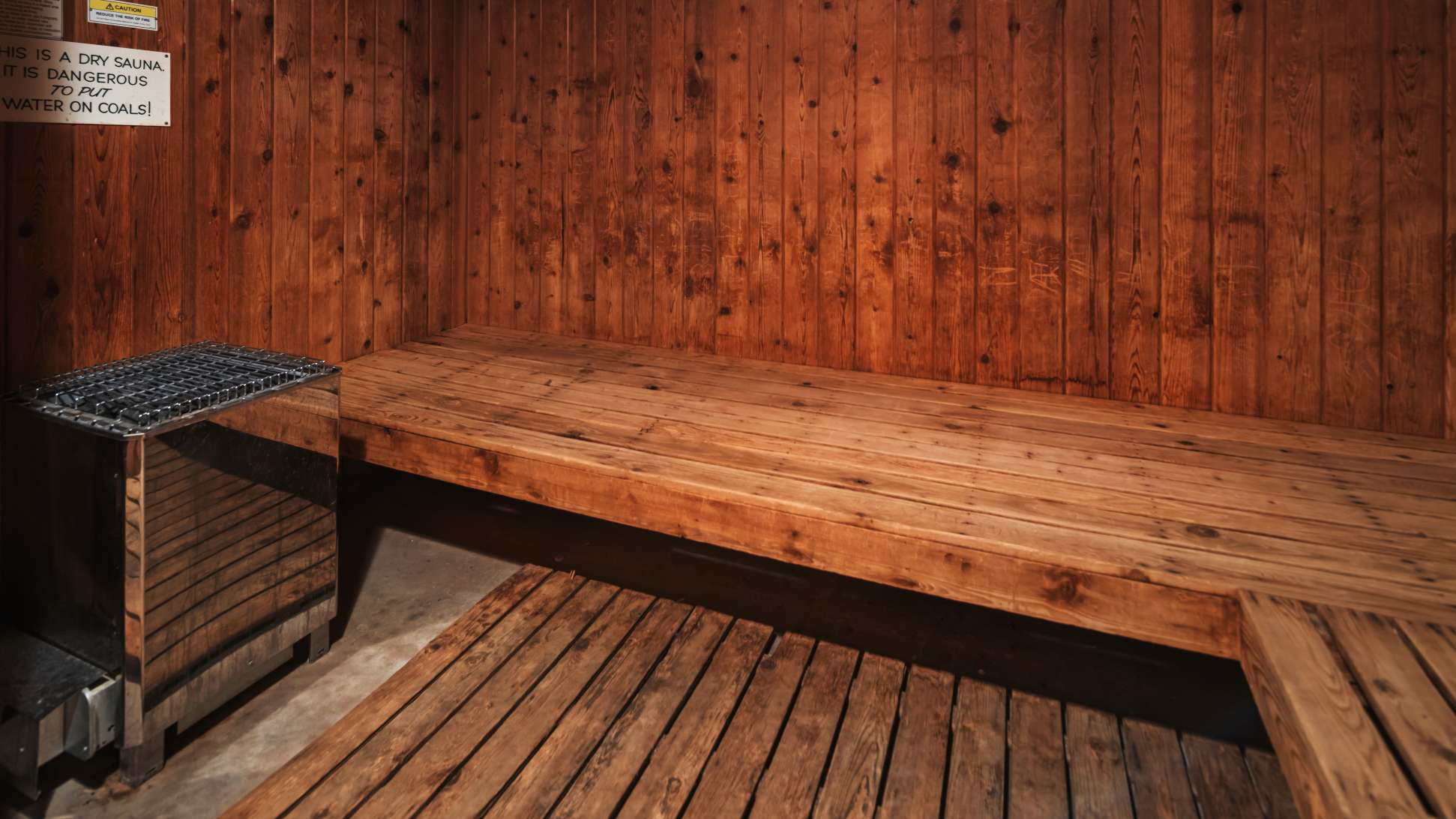 A rustic dry sauna features warm-toned wooden walls and benches, with a metal sauna heater visible on the left. A warning sign about not putting water on coals is posted on the wall.