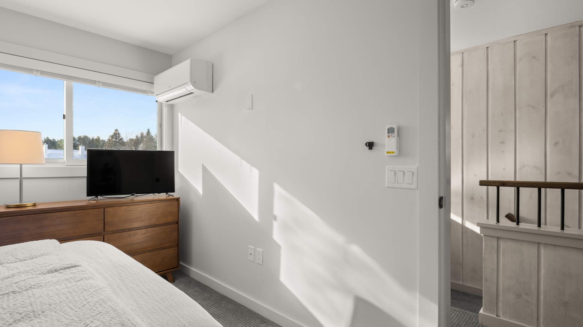 A corner of bedroom-1 showing a wooden dresser with a flat-screen TV, a large window overlooking a snowy landscape, and a wall-mounted AC unit.