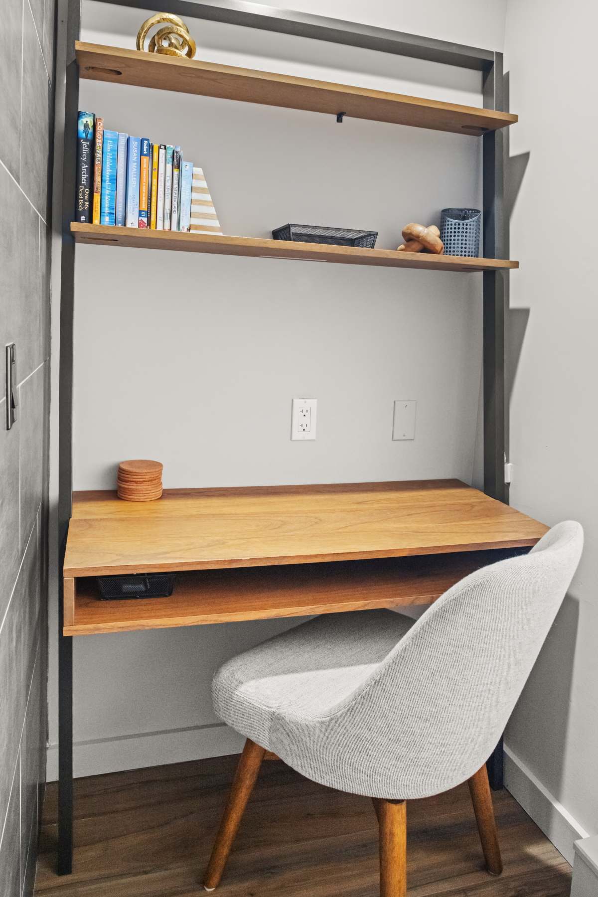 A compact and modern workspace features a light wood desk with a gray upholstered chair, built into a shelving unit. The wall is painted light gray, and the floor has wood-look tiles.