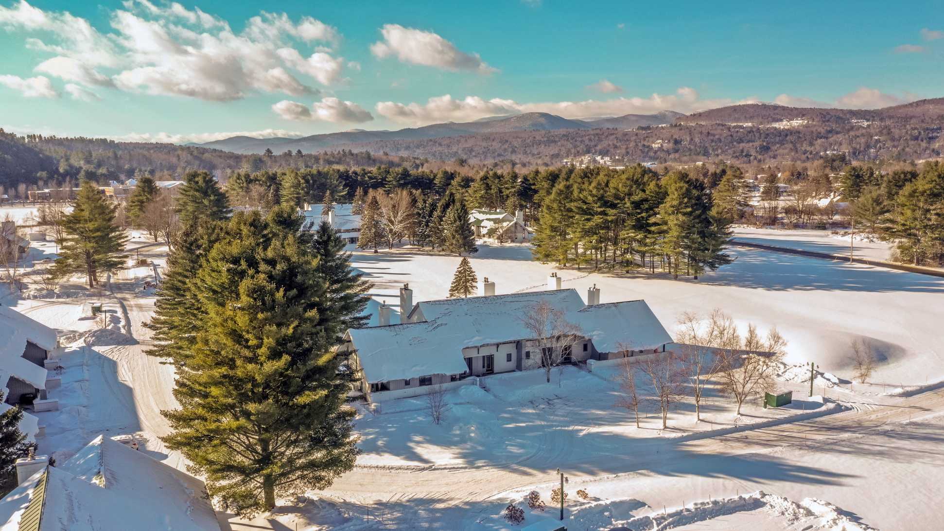 An aerial view captures a snow-covered condominium complex nestled among evergreen trees, with distant mountains under a partly cloudy sky. The white roofs contrast with the dark trees and blue sky.