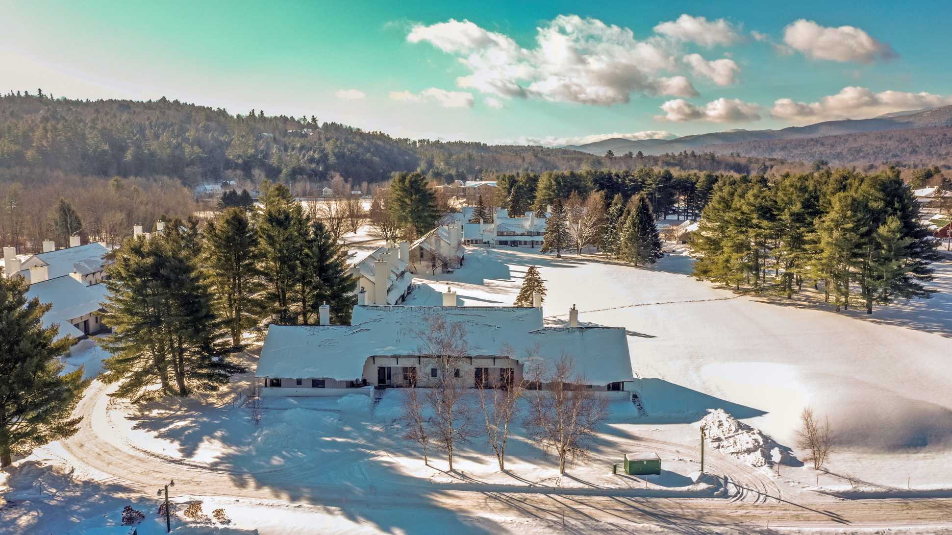 An aerial view of a snow-covered village with multiple buildings, surrounded by pine trees and rolling hills under a bright, cloudy sky, indicating a winter landscape.