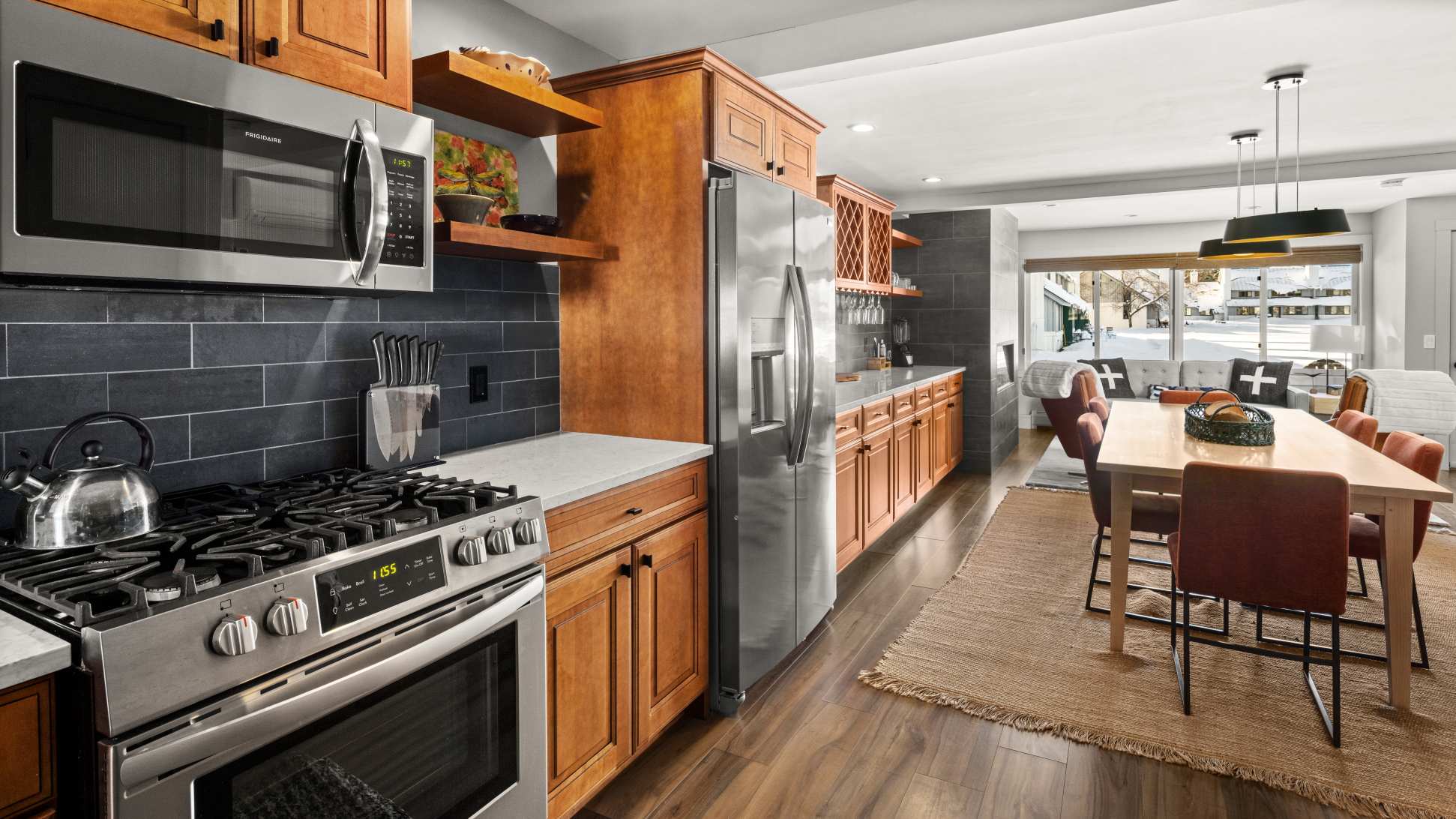 Open concept kitchen and dining area with light wood cabinetry, stainless steel appliances including a refrigerator and gas range, and a gray subway tile backsplash. A dining table with orange chairs is visible in the foreground.