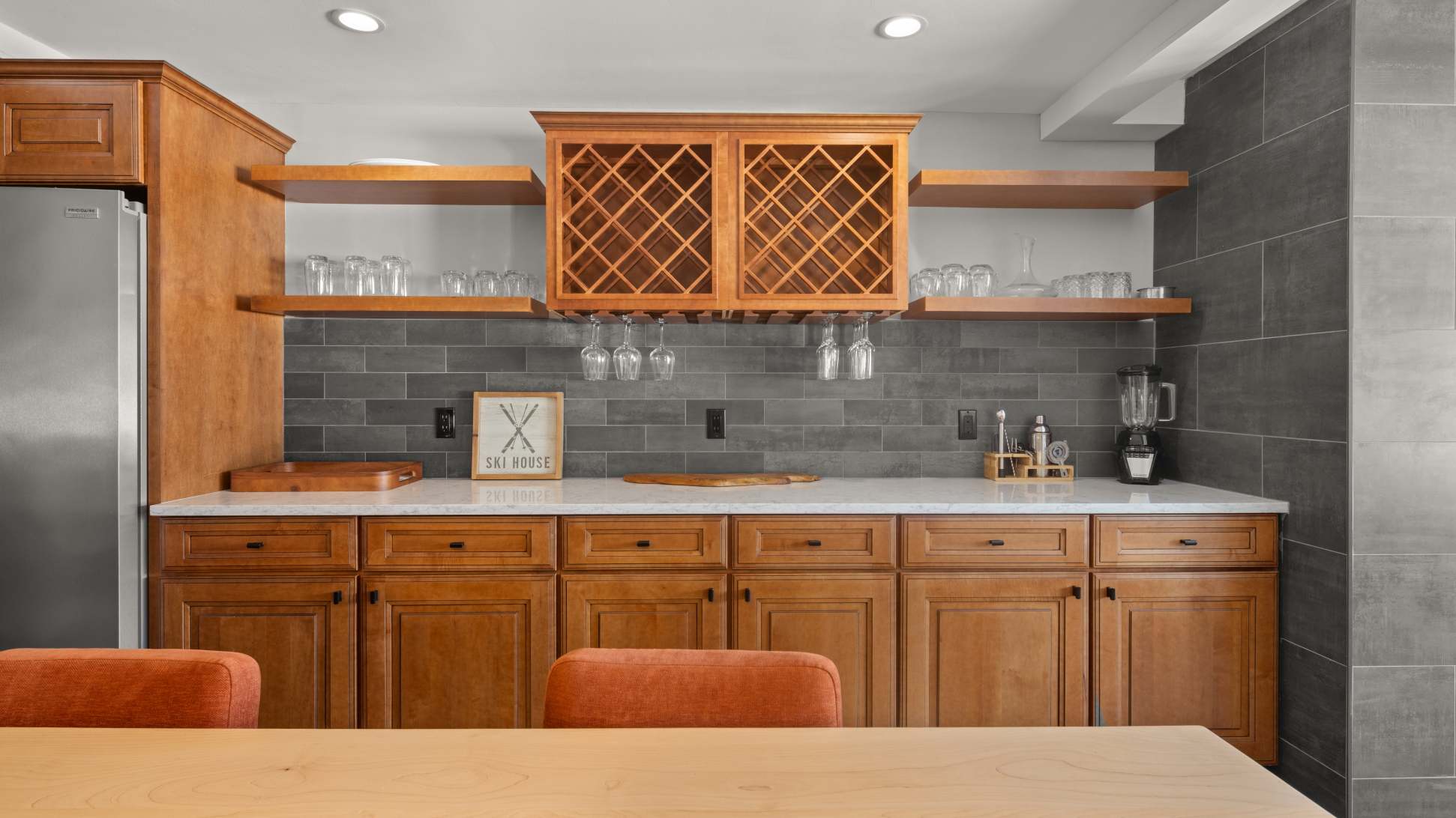 Wet bar area with light wood cabinetry, white countertops, a gray subway tile backsplash, and open shelving with glassware. A built-in wine rack is featured above.